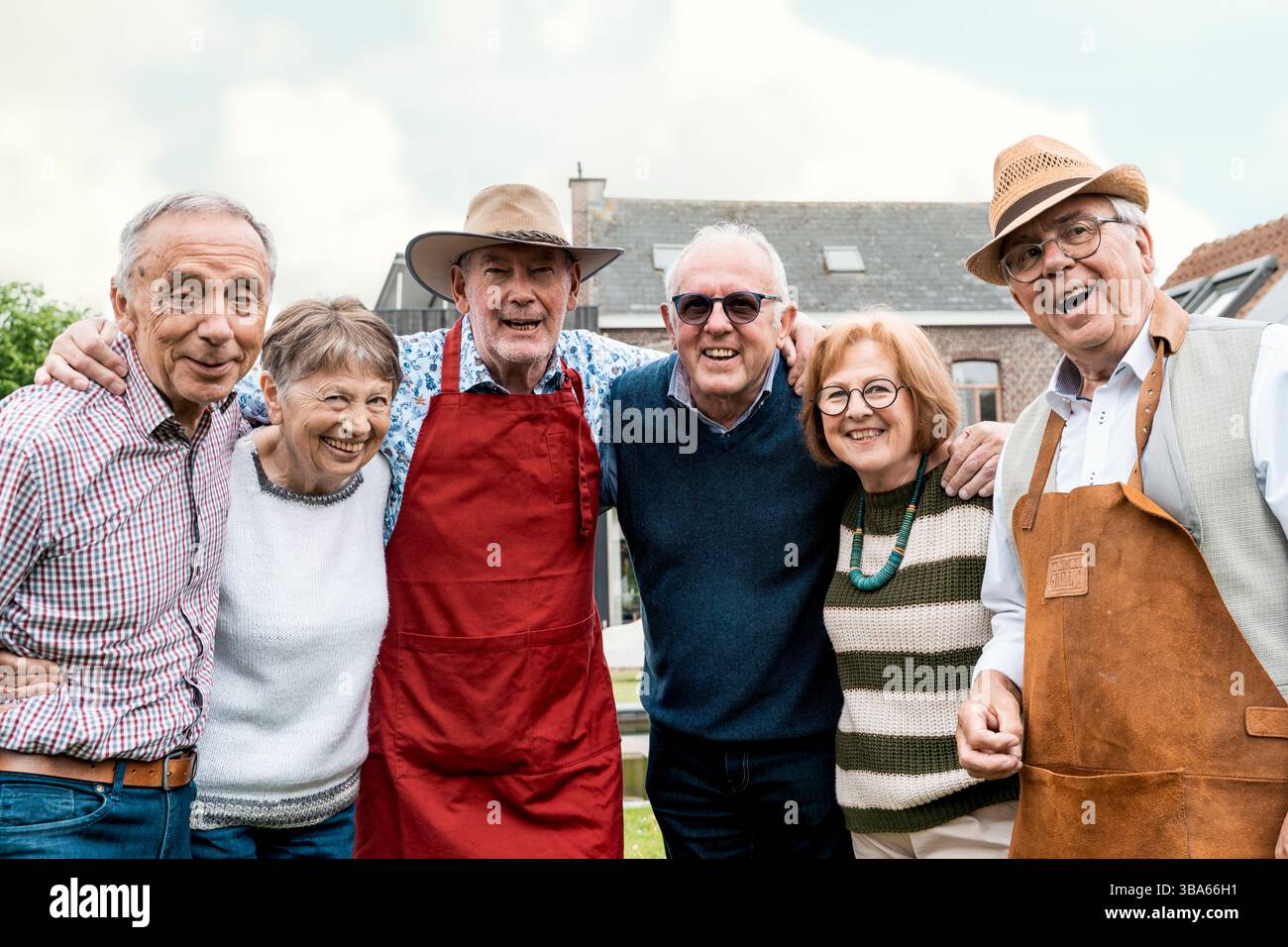 Sei allegri pensionati si alzano con un braccio in un giardino, sorridendo di fronte alla loro casa mentre indossano abiti casual e grembiuli grigliati, celebrando il frien Foto Stock
