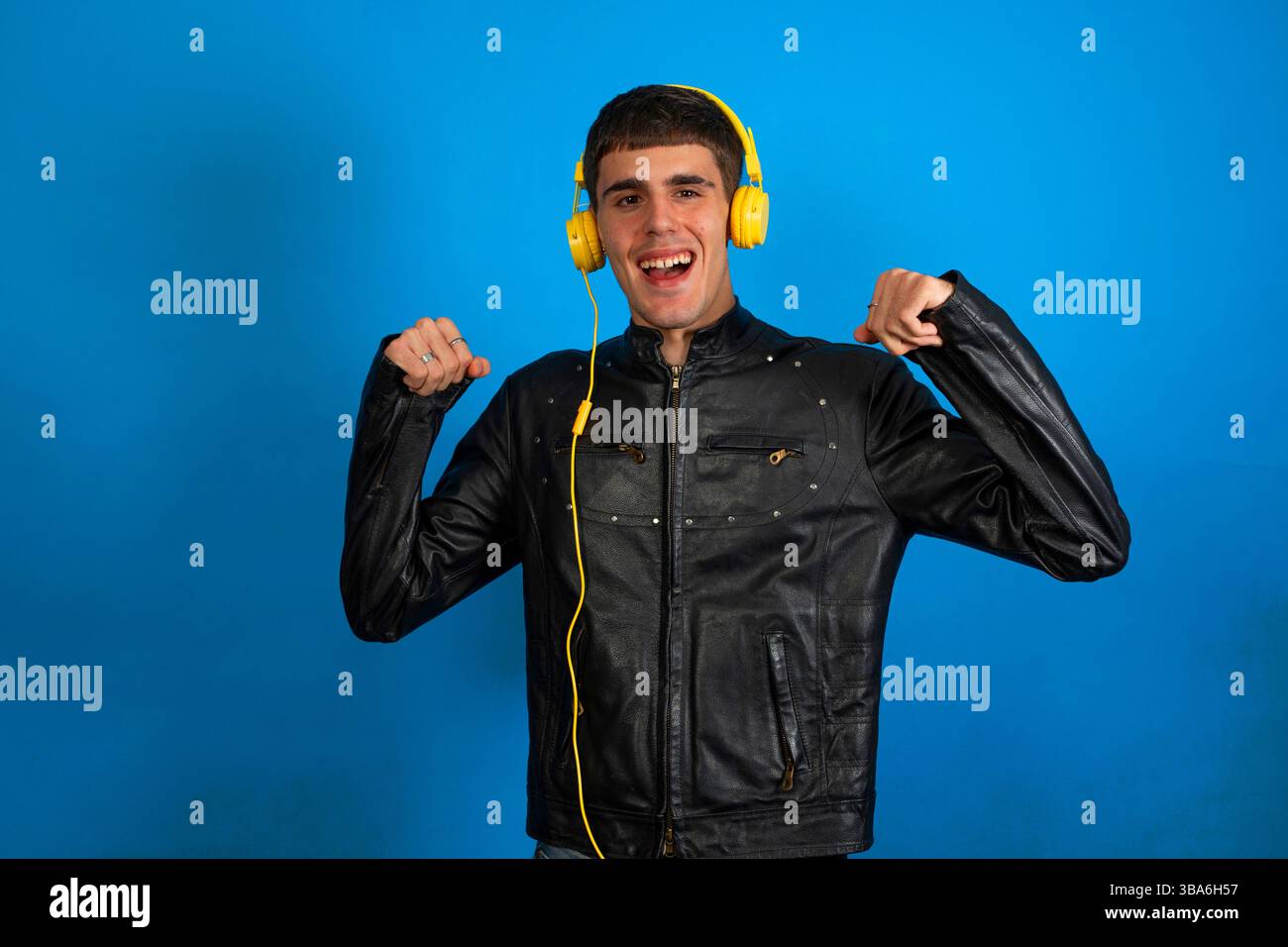 Uomo elegante che indossa cuffie gialle e giacca in pelle nera, che ascolta musica e balla su sfondo blu Foto Stock