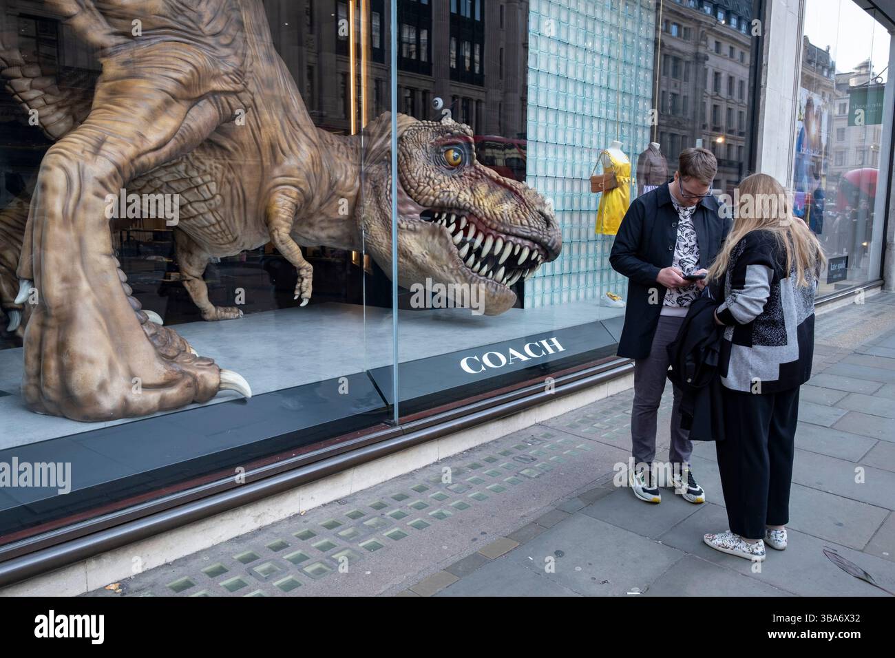 I visitatori di Regent Street passano davanti ad un modello in grande scala di un Tyrannosaurus Rex nella vetrina del negozio di Coach il 24 marzo 2025 a Londra, Regno Unito. Regent Street è un importante centro commerciale nel West End della capitale ed è conosciuta per i suoi negozi di punta, la maggior parte dei quali sono negozi di moda, di alta moda e di abbigliamento di fascia alta. Foto Stock