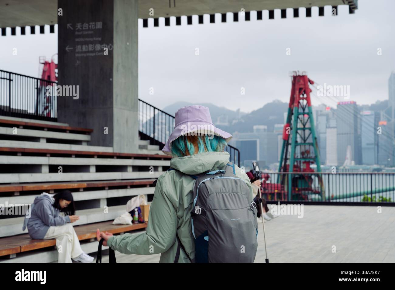Il visitatore con i suoi vivaci capelli blu e un cappello da secchio lilla passeggia davanti a una sezione di sbiancanti all'esterno del Museo M+ di Hong Kong. Foto Stock