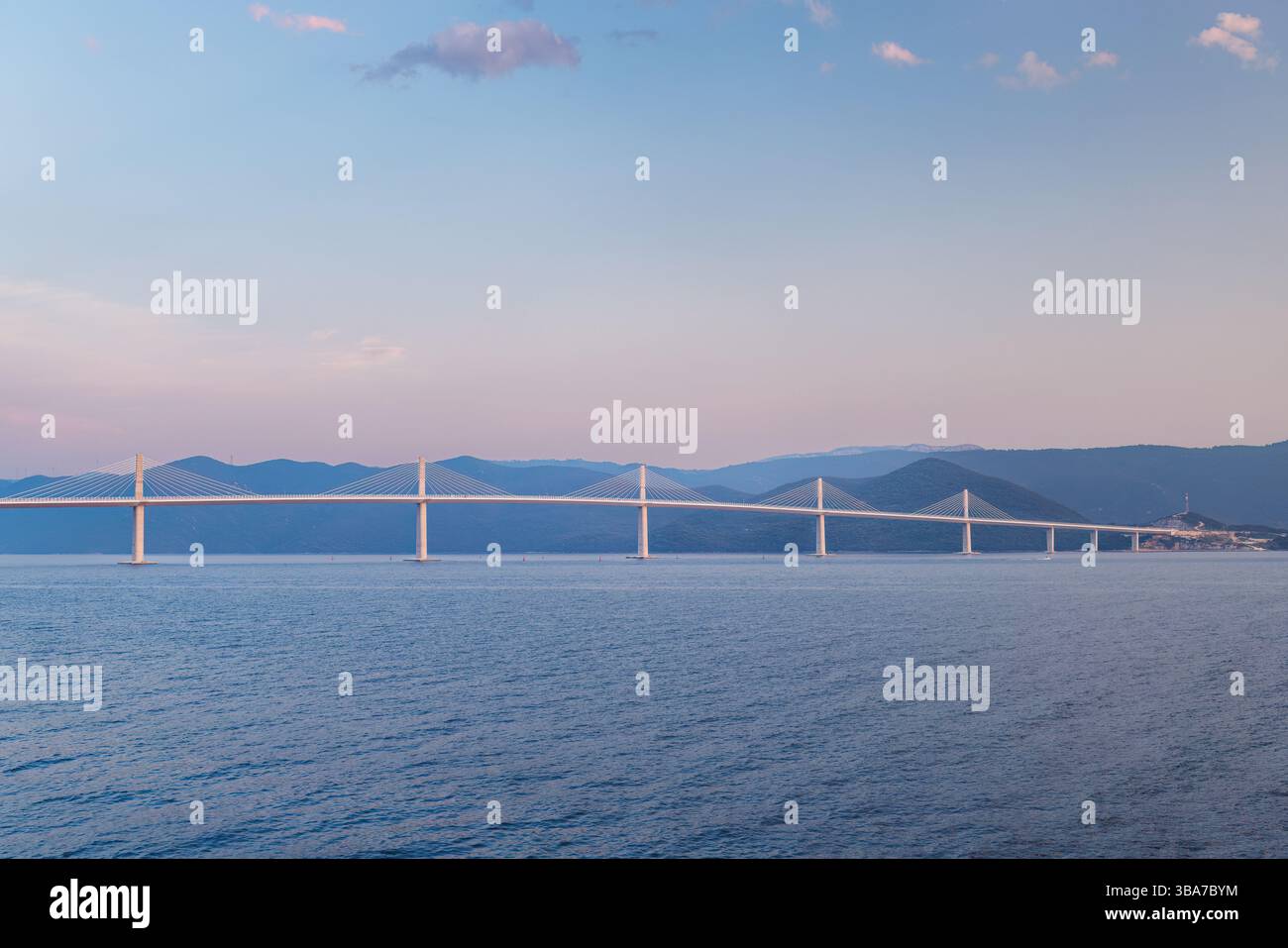 Un ponte lungo e moderno si estende su un'ampia distesa d'acqua, con dolci colline sullo sfondo sotto un cielo pastello. L'immagine evoca un senso di serenità trans Foto Stock