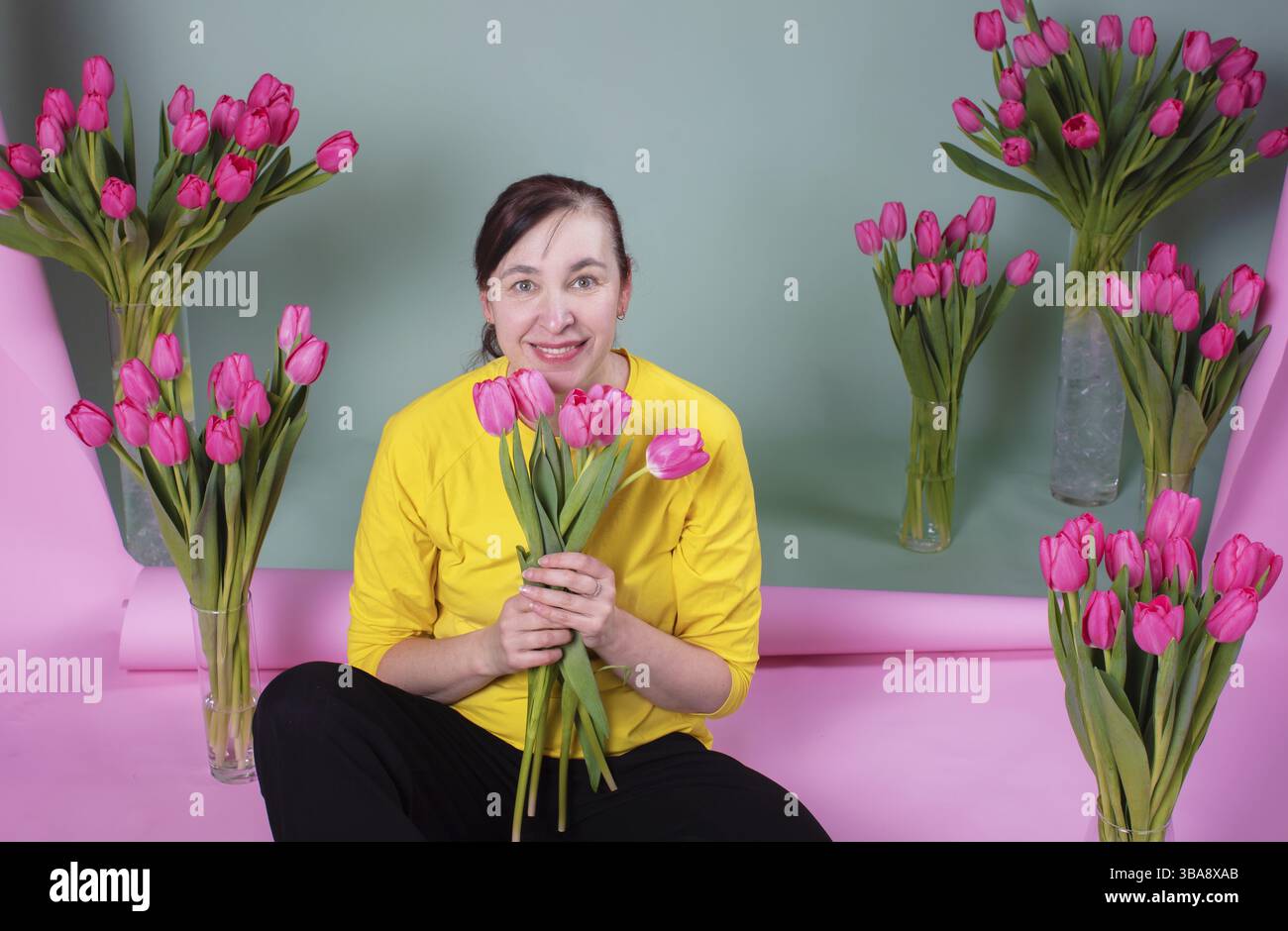 Una donna di mezza età felice con un mucchio di tulipani è felice e sorridente su uno sfondo rosa-verde Foto Stock