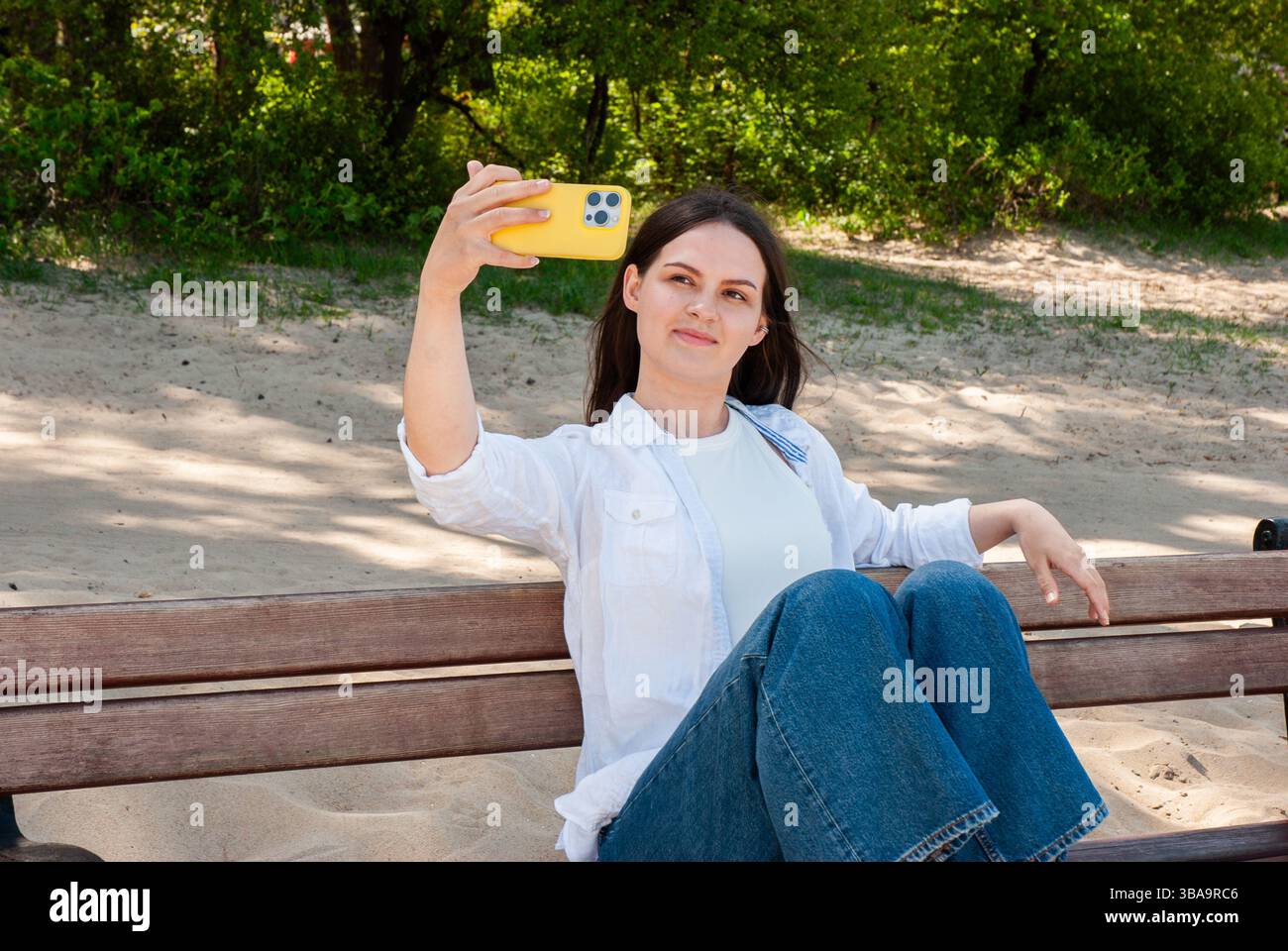 Giovane donna caucasica che scatta un selfie con lo smartphone giallo in una giornata di sole in spiaggia e si rilassa sulla panca di legno in un outfit casual. Nomad lif. Digitale Foto Stock