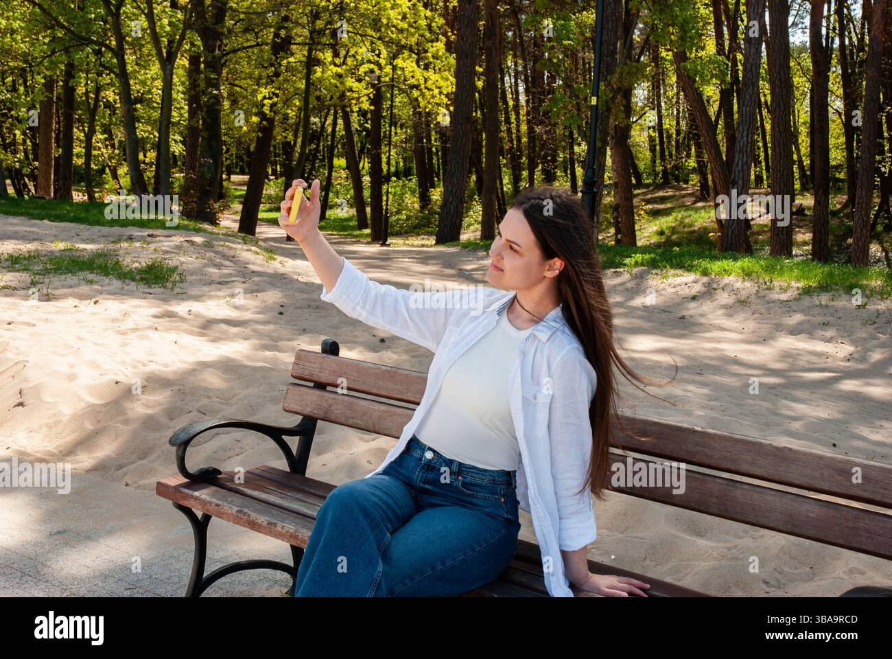 Giovane donna caucasica che scatta un selfie su una panchina di legno del parco circondata da lussureggianti alberi verdi e sentieri sabbiosi in una giornata di sole. Stile di vita digitale nomade. Foto Stock