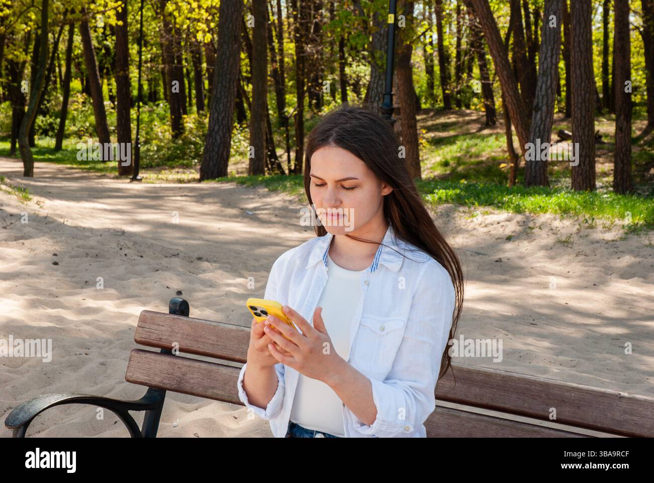 Giovane donna caucasica impegnata con lo smartphone in una giornata di sole sulla panchina del parco tra lussureggianti alberi verdi e sereno sentiero sabbioso in primavera. Nomade digitale Foto Stock