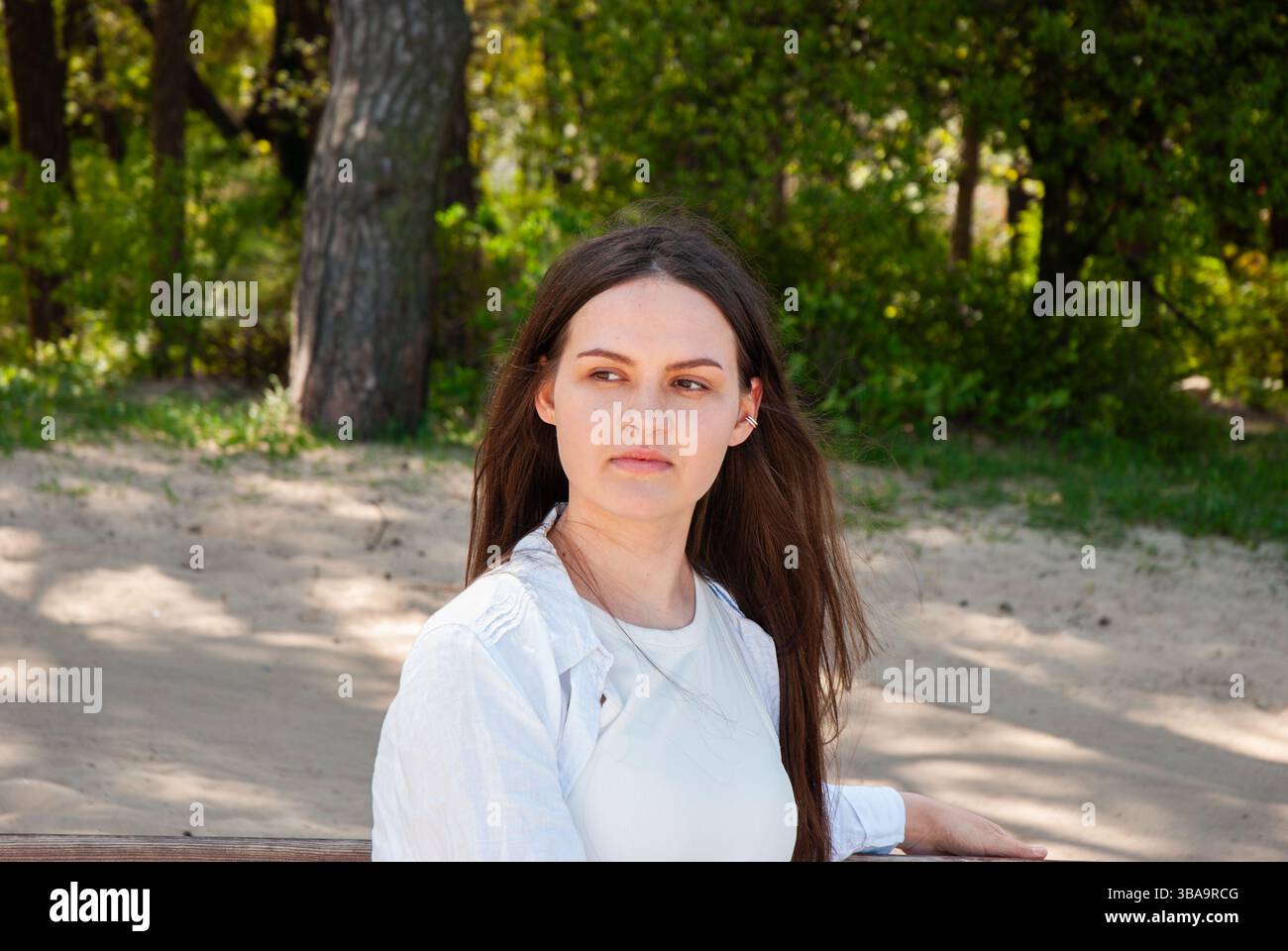 Giovane donna caucasica con lunghi capelli castani seduta su una panchina in una spiaggia di sabbia, circondata da lussureggianti alberi verdi in una giornata di sole. Nomade Lifestyl digitale Foto Stock