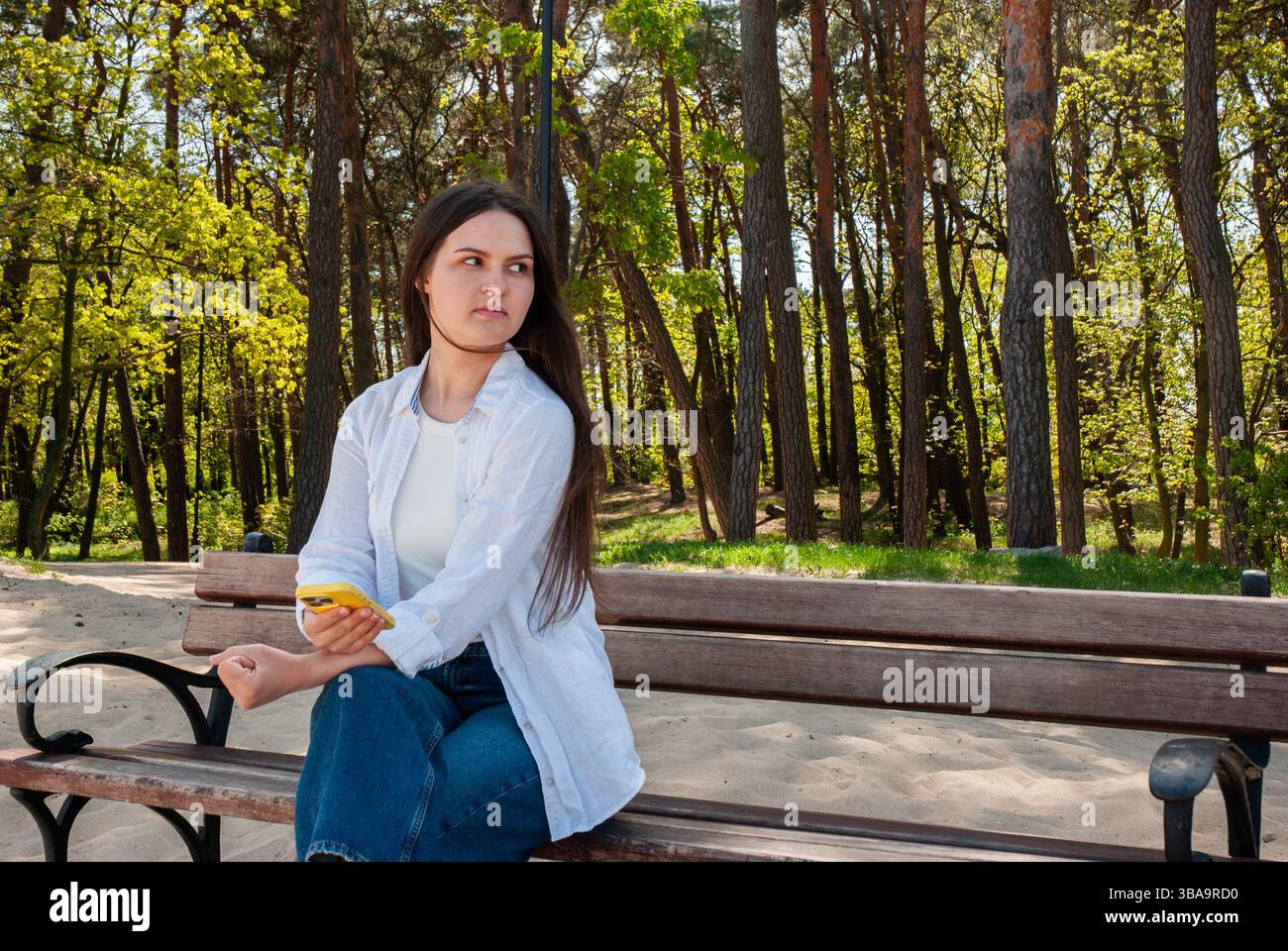 Giovane donna caucasica che si rilassa sulla panchina del parco nella foresta primaverile, tiene il telefono, indossa camicia bianca informale e jeans, con un aspetto premuroso. Nomade digitale Foto Stock