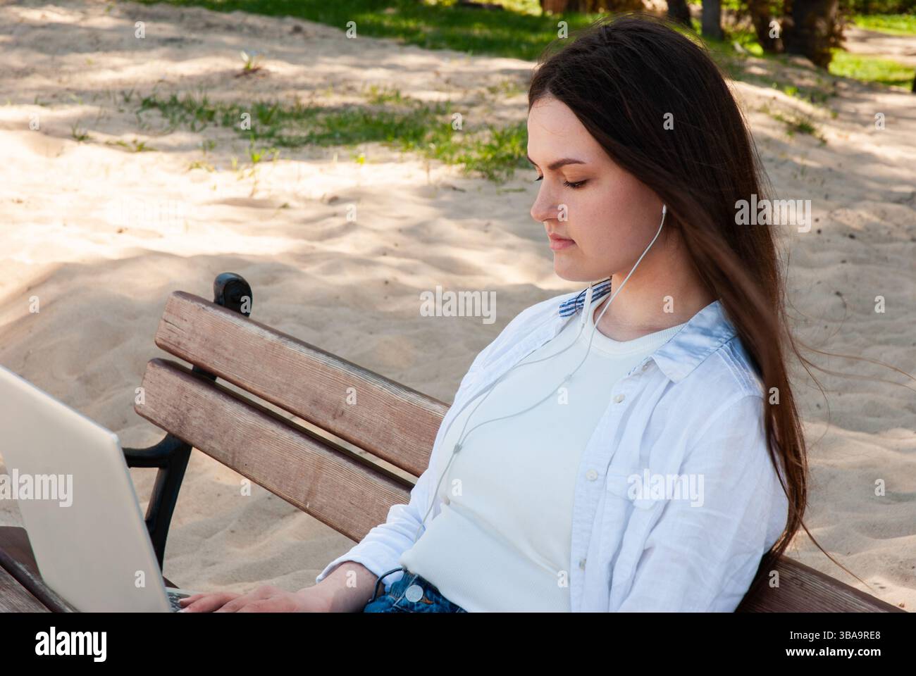 Giovane donna caucasica che si rilassa su una panchina del parco usando il computer portatile e le cuffie, godendosi una giornata di sole in un ambiente tranquillo e sabbioso. Nomade digitale Foto Stock