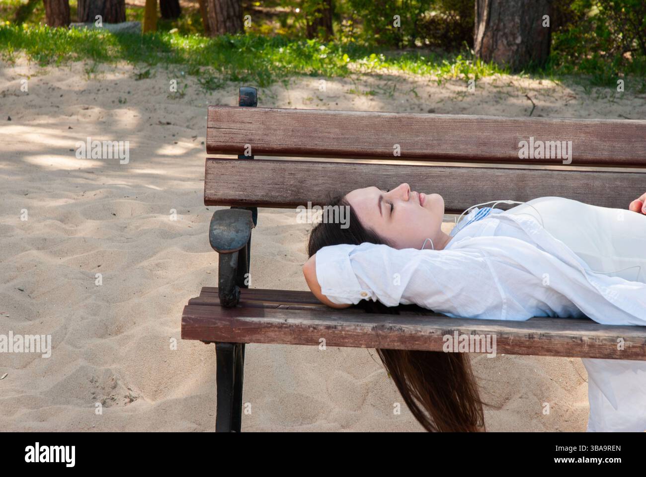 Giovane donna caucasica che si rilassa sulla panchina del parco di legno sulla spiaggia sabbiosa, godendosi un tranquillo pomeriggio estivo all'ombra degli alberi. Stile di vita dei nomadi digitali Foto Stock