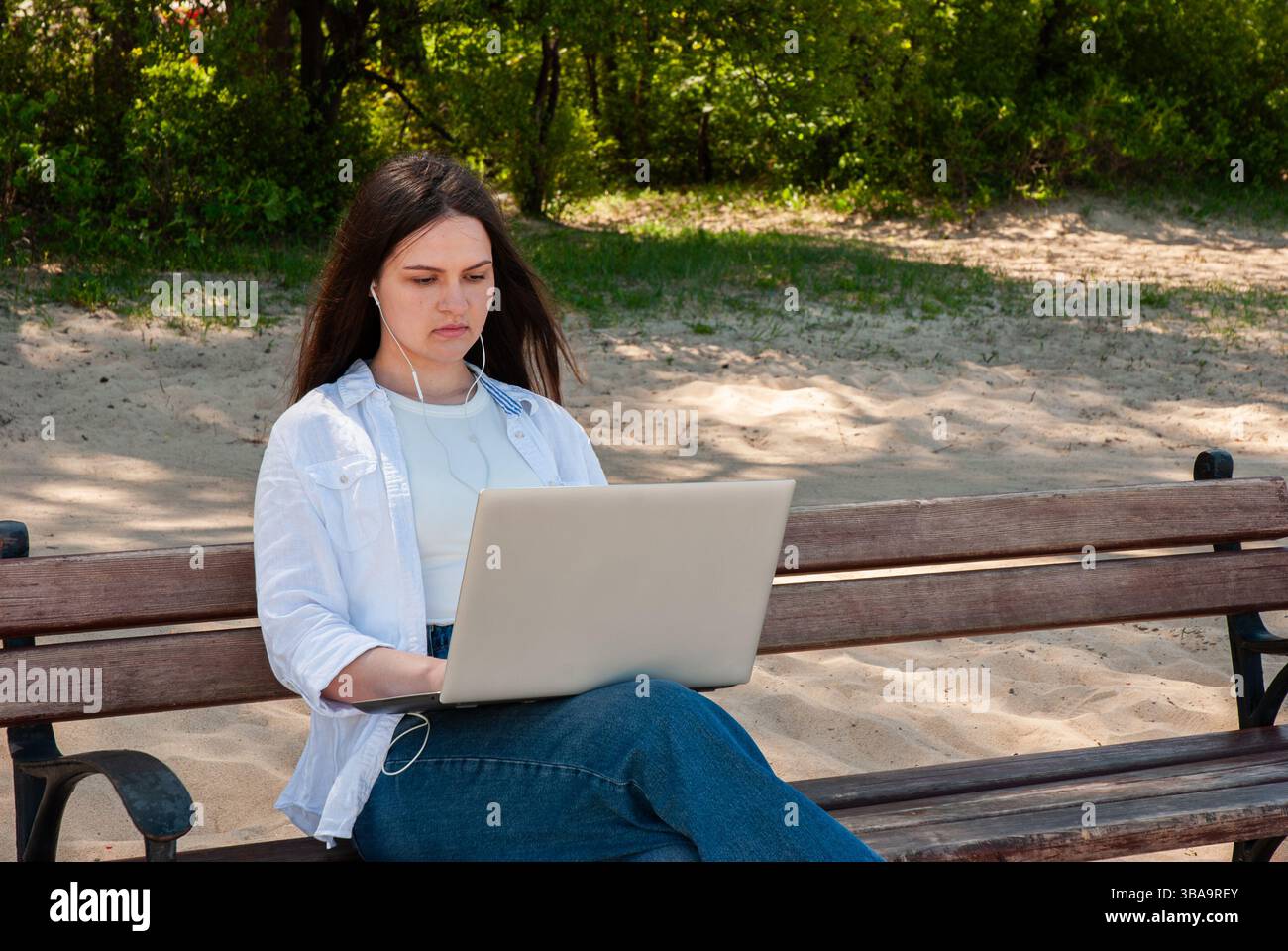 Giovane donna caucasica che usa un computer portatile con auricolari sulla panca del parco nelle giornate di sole, concentrata sul lavoro e godendo della tranquillità della natura. Il lifest dei nomadi digitali Foto Stock