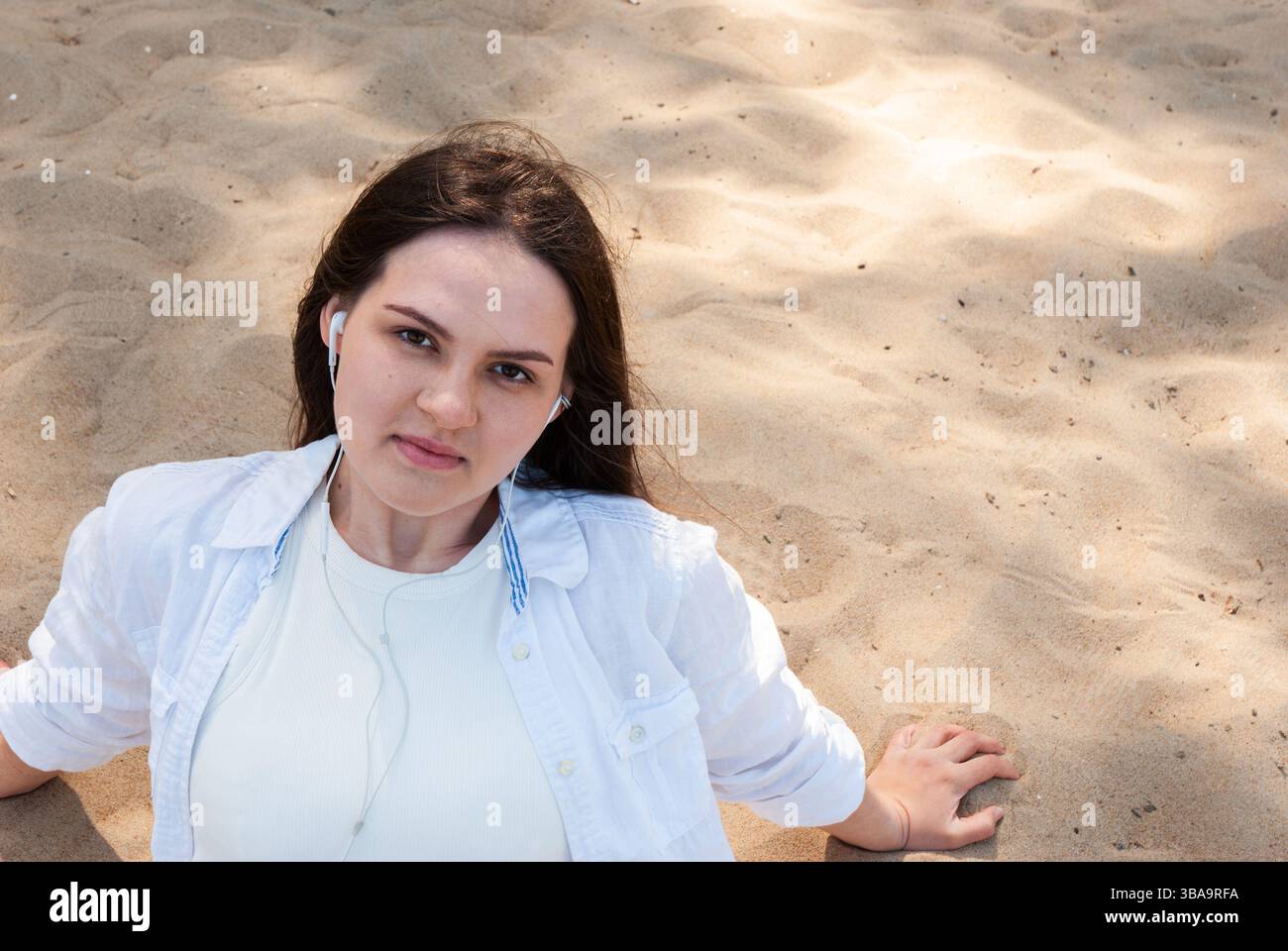 Giovane donna caucasica che si rilassa sulla spiaggia di sabbia ascoltando musica all'aperto con auricolari in una giornata di sole in camicia bianca informale e in un ambiente naturale. Di Foto Stock