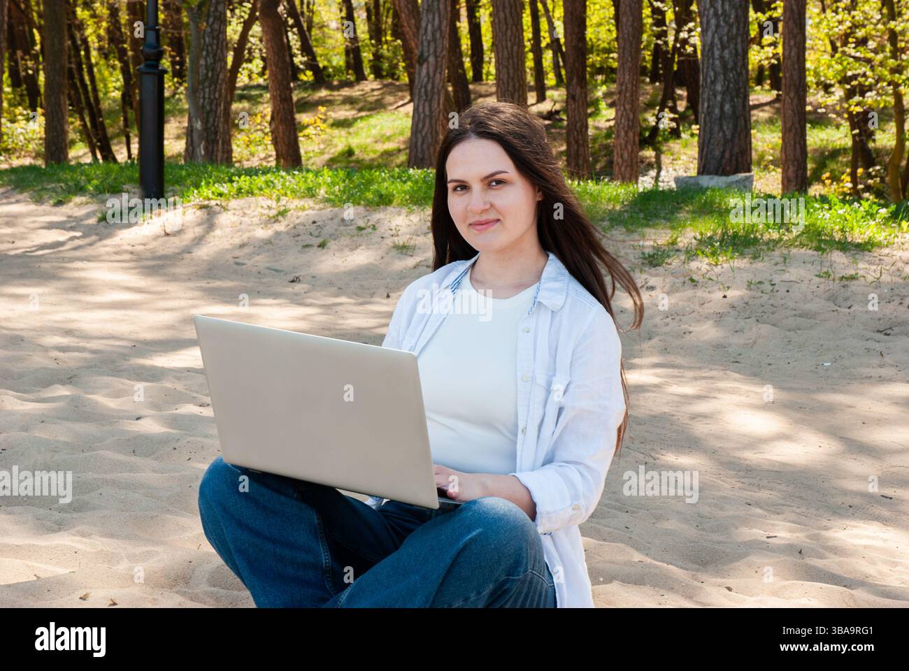 Giovane donna caucasica con capelli lunghi seduta sulla spiaggia di sabbia usando un laptop in abiti informali circondata da lussureggianti boschi. Nomad digitale Foto Stock