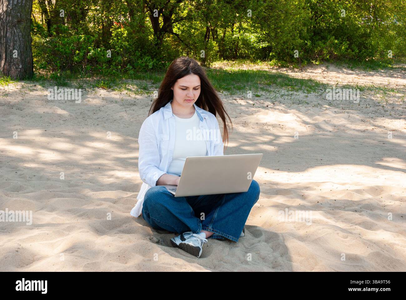 La giovane donna caucasica con un abbigliamento casual si concentra sul laptop mentre si siede su una spiaggia di sabbia assolata all'ombra di lussureggianti alberi verdi. Nomade digitale Foto Stock
