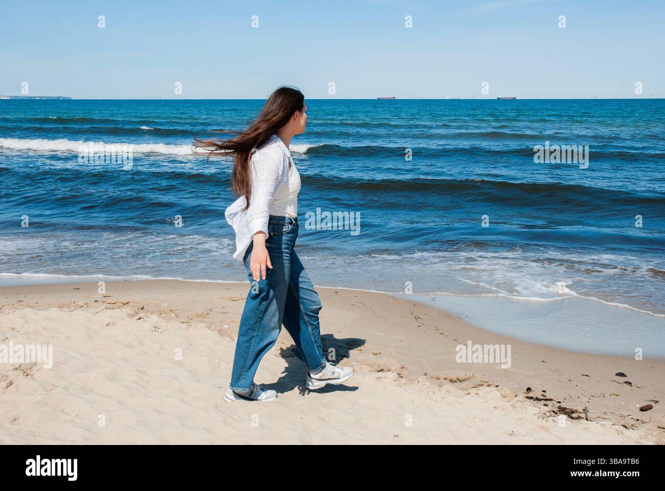 La giovane donna asiatica cammina lungo la spiaggia sabbiosa sotto un cielo azzurro limpido con dolci onde sullo sfondo, godendosi una tranquilla giornata al mare. Digitale no Foto Stock
