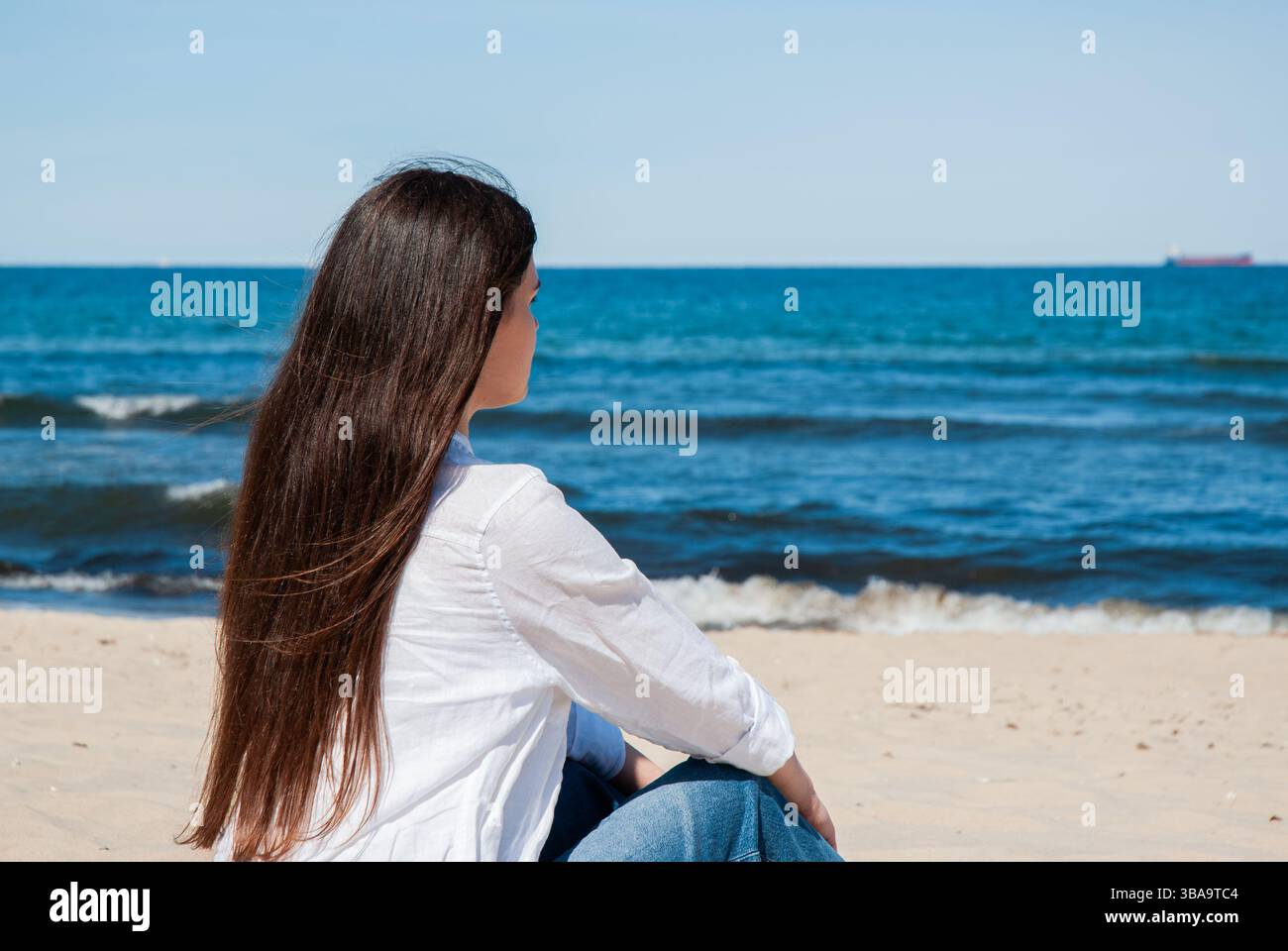 Giovane donna caucasica seduto su una spiaggia sabbiosa che guarda il tranquillo oceano blu sotto un cielo limpido, godendosi un tranquillo momento di mare. Digitale Foto Stock