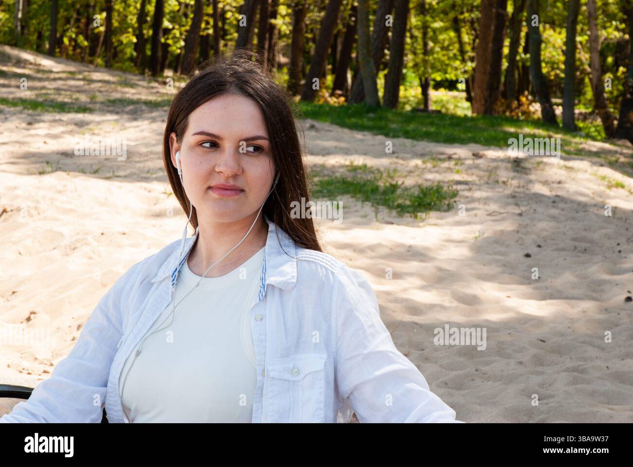 Giovane donna caucasica che ascolta la musica attraverso le cuffie mentre siede su una spiaggia sabbiosa circondata da lussureggianti alberi verdi in una giornata di sole. Nomad digitale li Foto Stock
