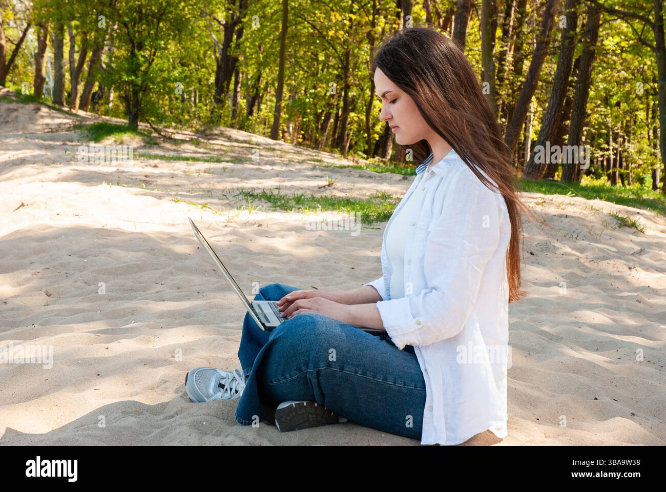 Giovane donna caucasica con capelli lunghi che usa un laptop all'aperto su una spiaggia sabbiosa circondata da alberi verdi in una brillante giornata primaverile. Stile di vita dei nomadi digitali Foto Stock