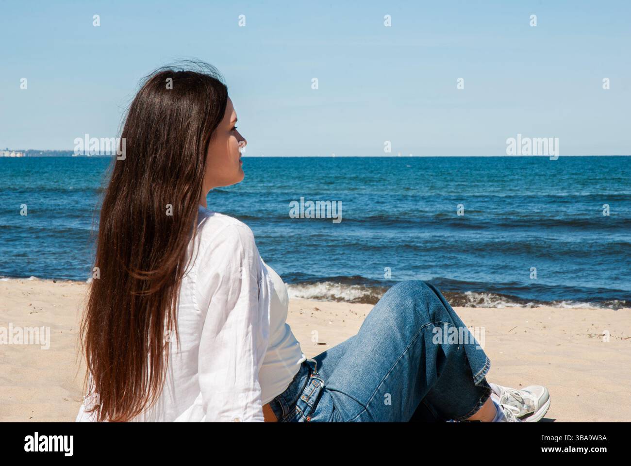 Giovane donna caucasica che si rilassa su una spiaggia soleggiata in un abbigliamento informale, godendo della vista sull'oceano e del cielo blu in tranquillità. Stile di vita dei nomadi digitali Foto Stock