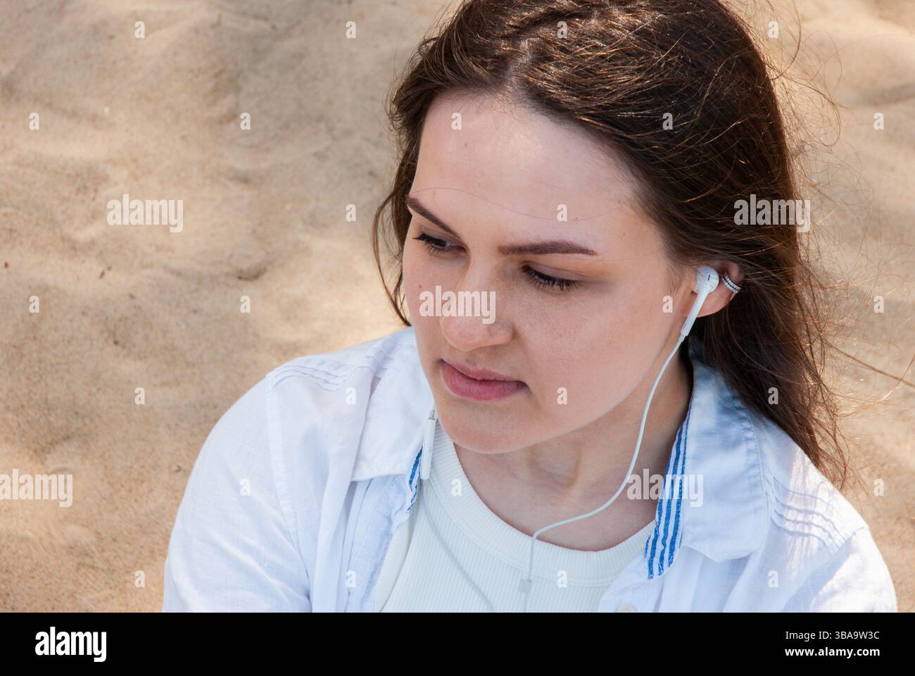 Giovane donna caucasica con lunghi capelli marroni che ascolta la musica sugli auricolari mentre siede sulla spiaggia di sabbia indossando una camicia bianca senza sorriso. Digitale Foto Stock