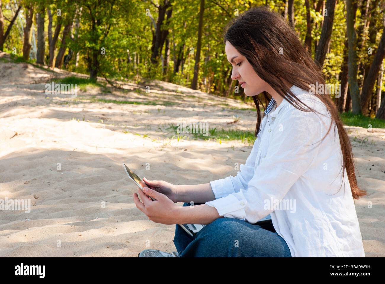 Giovane donna caucasica seduta su un sentiero di foresta sabbiosa leggendo un libro all'aperto in un abbigliamento casual sotto un vivace fogliame verde. Stile di vita digitale nomade. REM Foto Stock