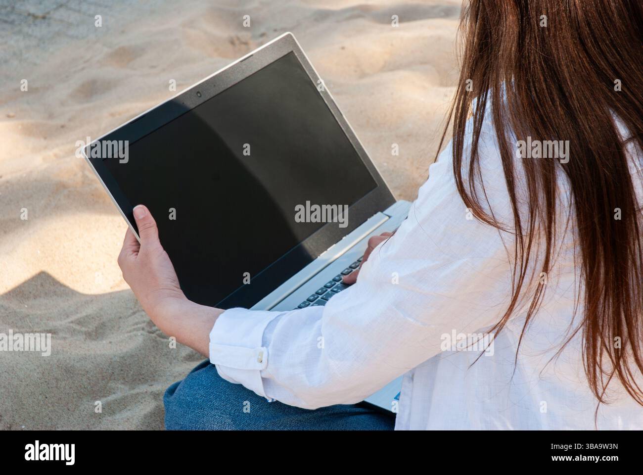 Giovane donna che lavora a distanza su un laptop mentre si siede sulla spiaggia di sabbia, abbraccia il sole estivo e mescola lavoro con relax in un ambiente informale Foto Stock