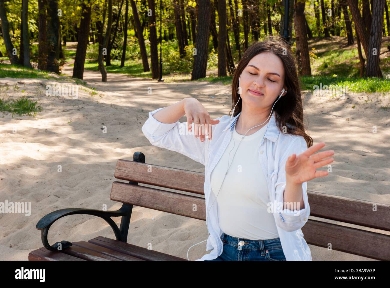 Giovane donna caucasica che ascolta musica in una giornata di sole, rilassarsi nella natura con le cuffie in un parco circondato da alberi e sabbia. Stile di vita dei nomadi digitali Foto Stock