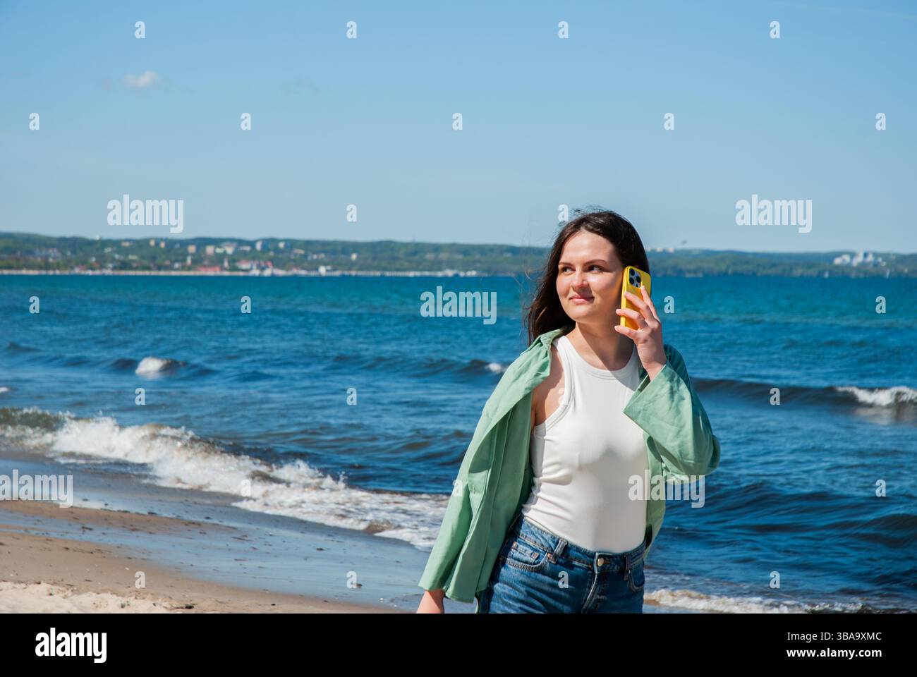 Giovane donna caucasica che cammina lungo la spiaggia con un telefono giallo in mano contro un cielo blu e sullo sfondo dell'oceano godendosi una giornata di sole. Nomad digitale Foto Stock