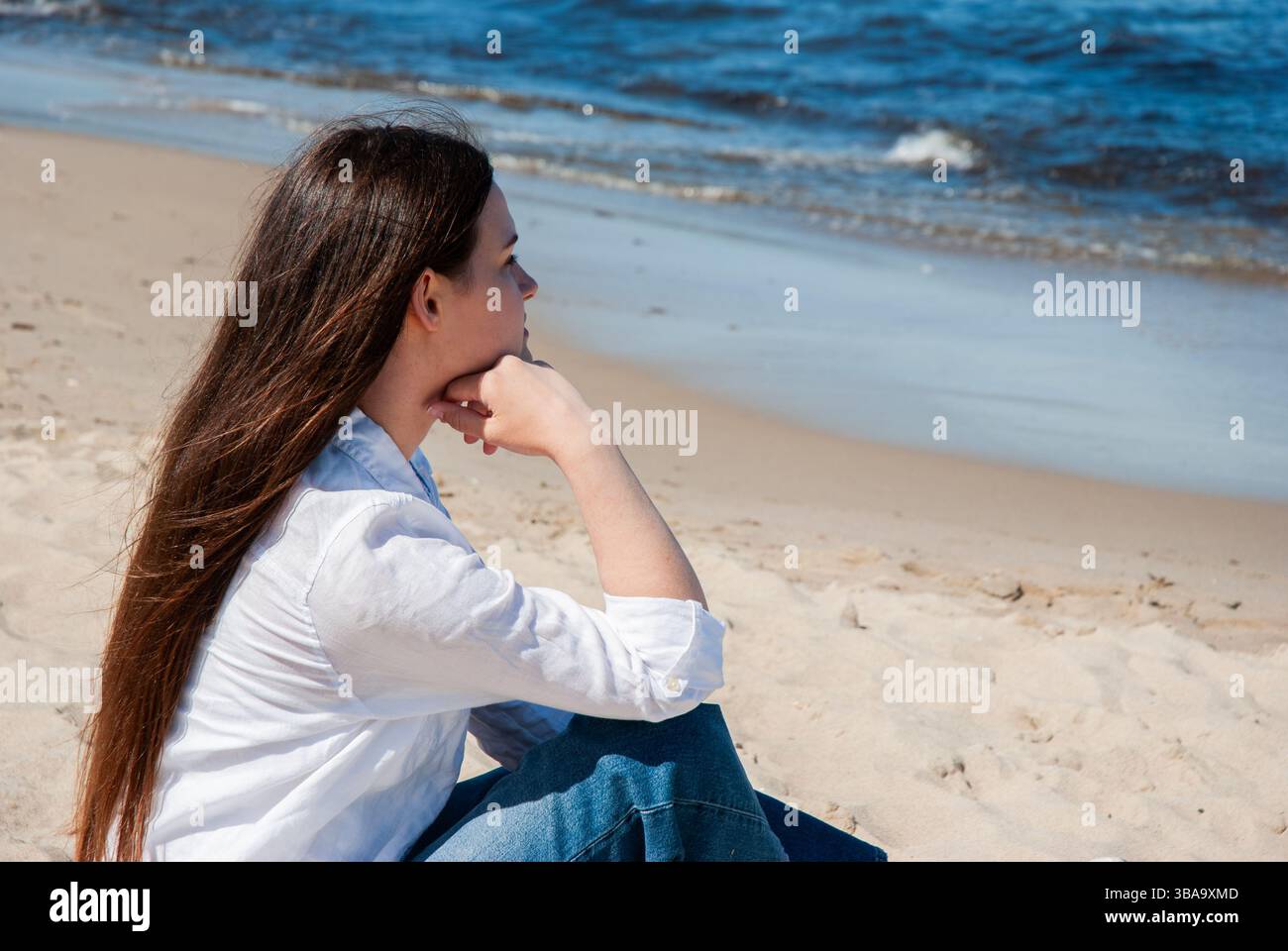 Giovane donna caucasica in camicia bianca e jeans siede con cura su una spiaggia di sabbia guardando attraverso le onde sotto un cielo azzurro e cristallino, abbracciando la tranquillità. Foto Stock