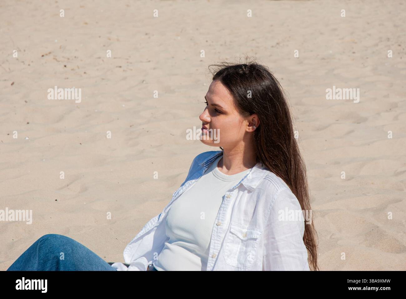 Giovane donna caucasica che si rilassa su una spiaggia soleggiata indossando una camicia bianca e jeans blu, godendosi il calore e la tranquillità di una giornata estiva. Digitale no Foto Stock