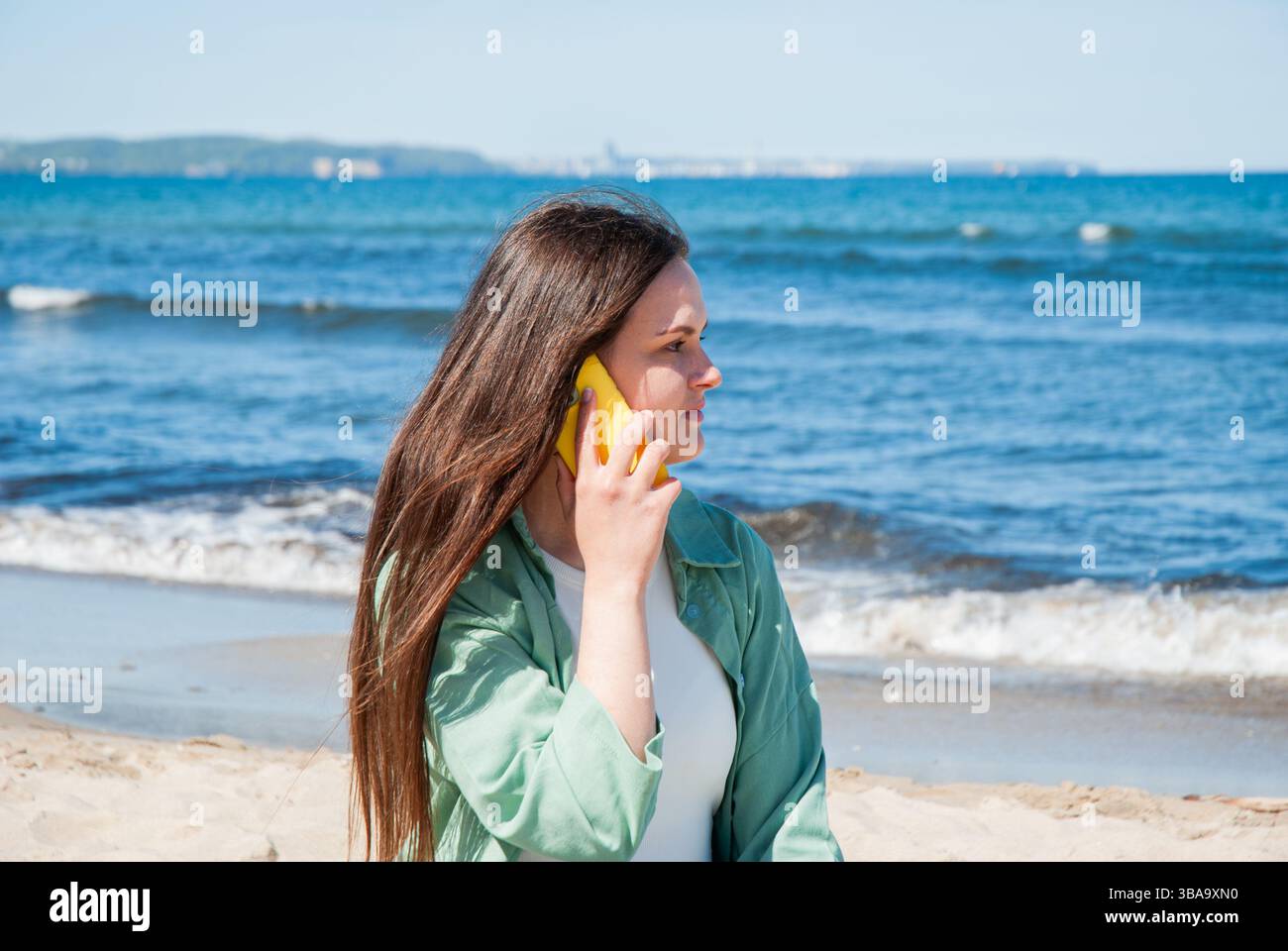 Giovane donna caucasica con lunghi capelli castani che parla su un telefono giallo in riva al mare, godendosi una giornata di sole sulla spiaggia sabbiosa con le onde sullo sfondo Foto Stock