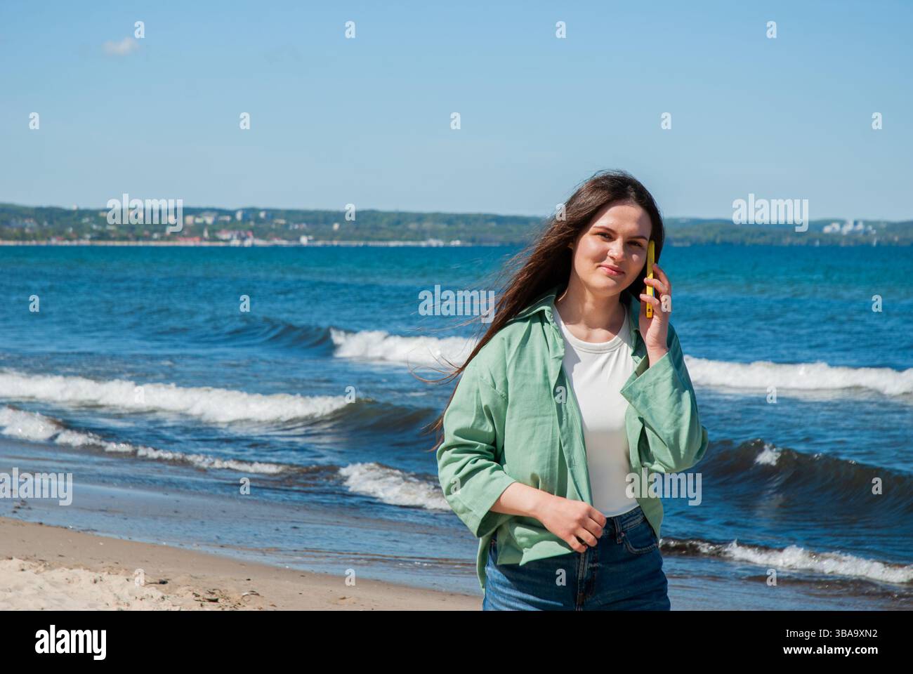 La giovane donna caucasica con i capelli lunghi parla al telefono sulla spiaggia soleggiata, indossa una giacca verde e un jeans, godendosi le onde con un cielo azzurro. Digi Foto Stock