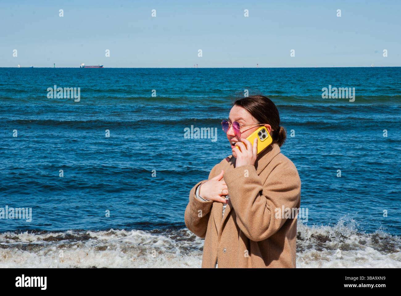 Giovane donna caucasica in spiaggia con indosso un cappotto marrone, che parla su un telefono giallo con occhiali da sole viola, sta in piedi in riva all'oceano in una giornata limpida. Digita Foto Stock