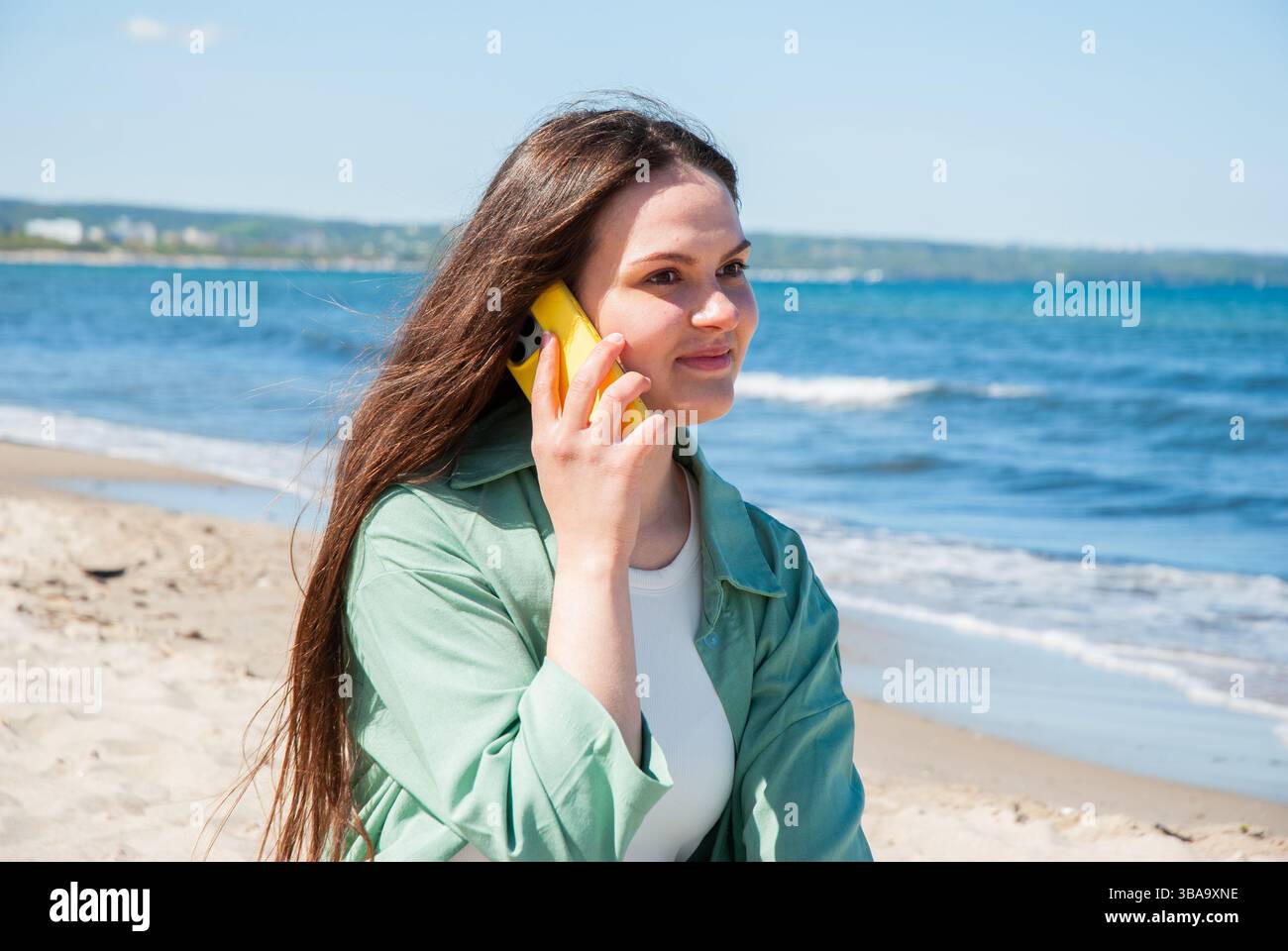 Giovane donna caucasica che si gode una giornata di sole sulla spiaggia, chiacchierando su uno smartphone giallo con un sorriso, onde marine turchesi e cielo blu sullo sfondo. D Foto Stock
