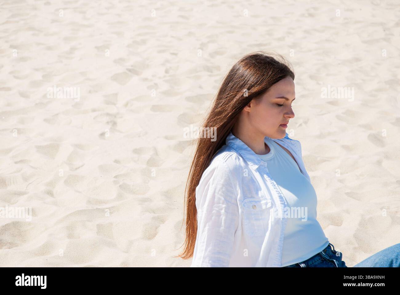 Giovane donna caucasica che si rilassa sulla spiaggia soleggiata con camicia bianca e jeans, circondata dalla sabbia sotto un cielo illuminato dal sole, con gli occhi chiusi per appagamento. Cifra Foto Stock