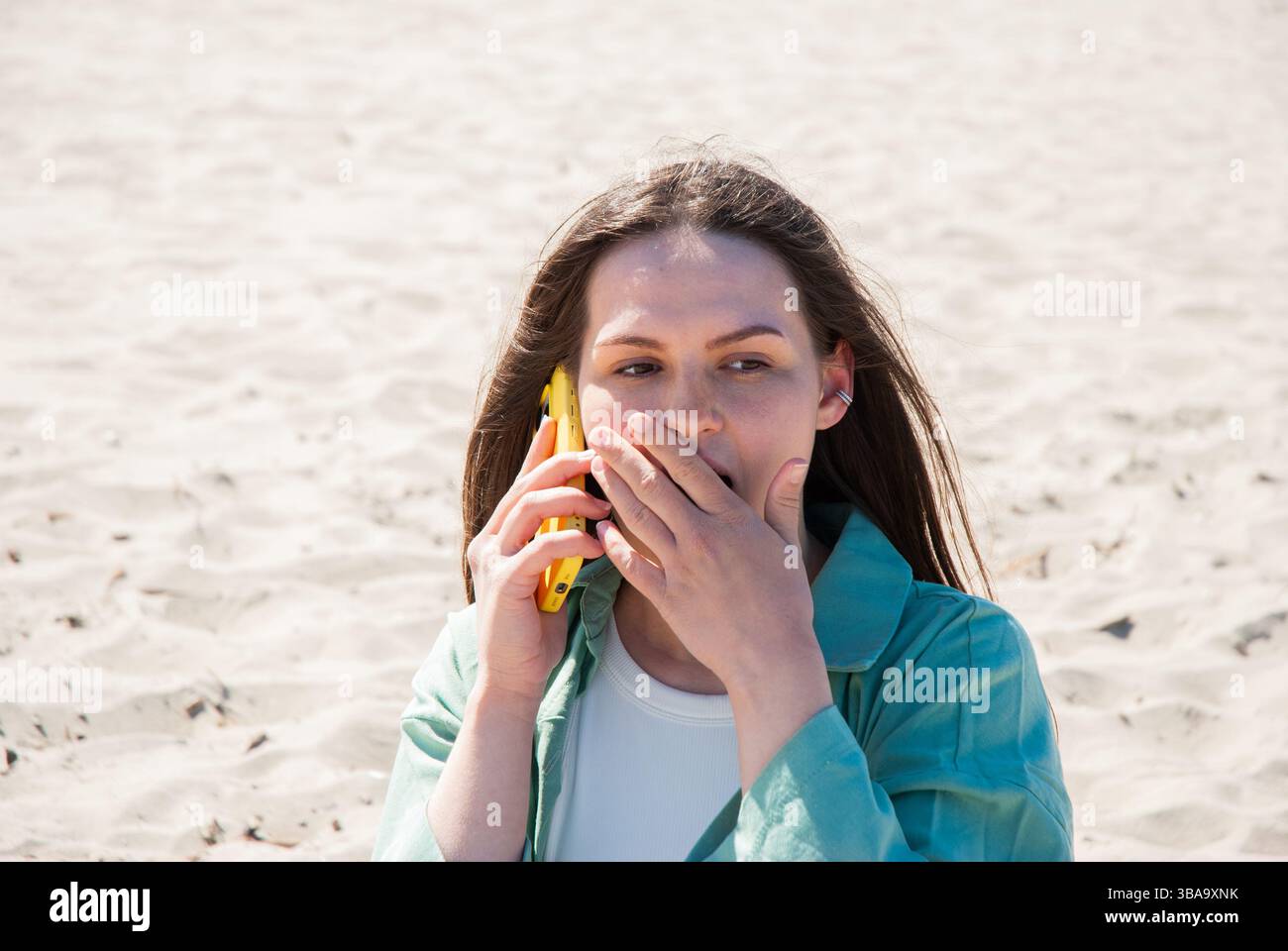 Giovane donna caucasica che parla al telefono giallo in un ambiente da spiaggia con espressioni sorprendenti e capelli battuti dal vento sotto la luce del sole. Nomad lif. Digitale Foto Stock