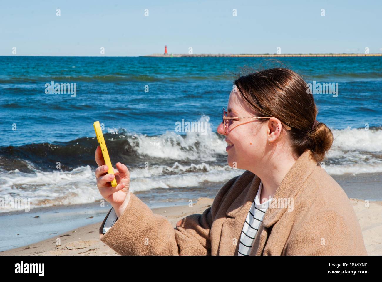 Giovane donna caucasica che si gode una rilassante giornata in spiaggia, sorridendo mentre scatta un selfie con il suo telefono sullo sfondo di onde e un lontano schiarito Foto Stock