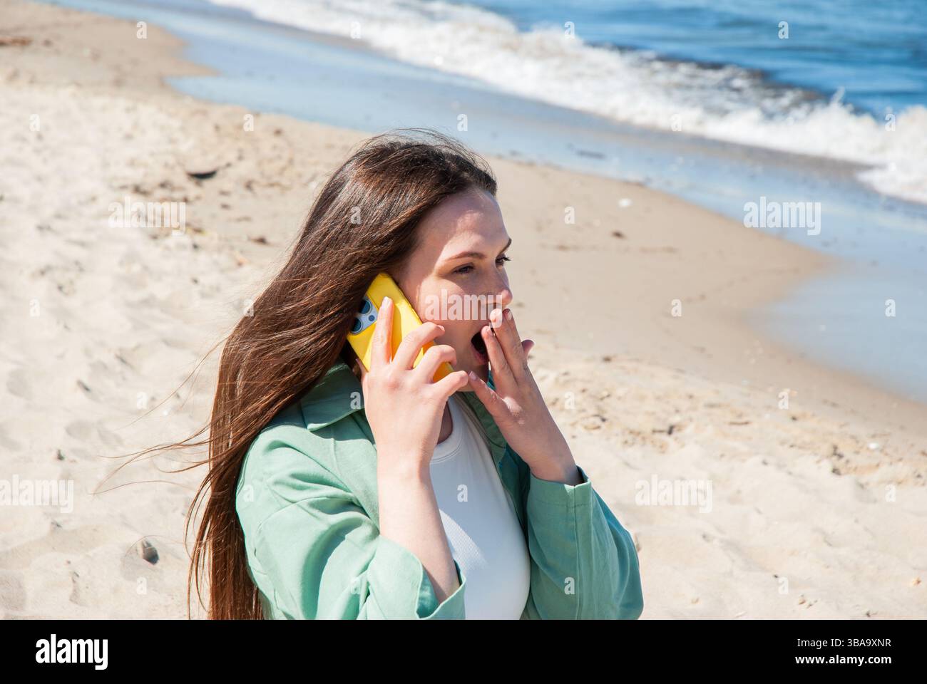 Giovane donna caucasica sulla spiaggia soleggiata che fa una telefonata a sorpresa con uno smartphone giallo brillante mentre le onde dell'oceano lambiscono dolcemente la riva. Nomade digitale Foto Stock