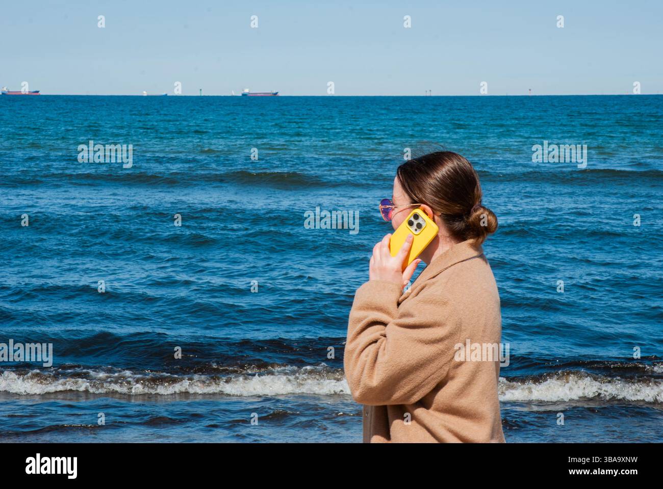Donna caucasica adulta che utilizza uno smartphone giallo sulla spiaggia di sabbia, indossa occhiali da sole e cappotto beige, di fronte alle calme onde blu dell'oceano. Vita nomade digitale Foto Stock