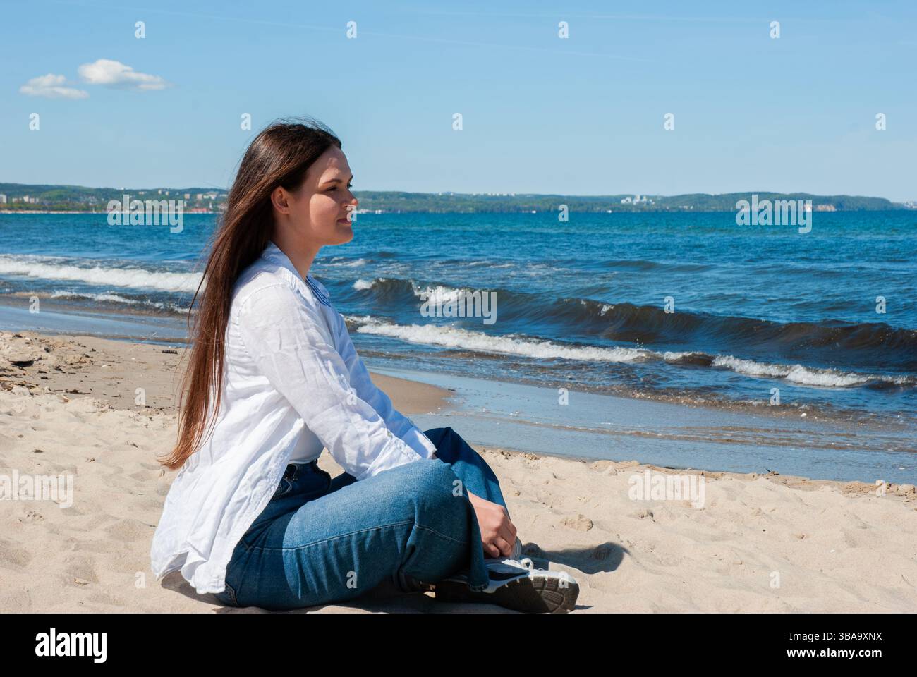 La giovane donna caucasica con un abbigliamento informale gode di un momento di serenità su una spiaggia soleggiata, circondata da onde blu e da una lontana vista sulla costa. Nomade digitale Foto Stock