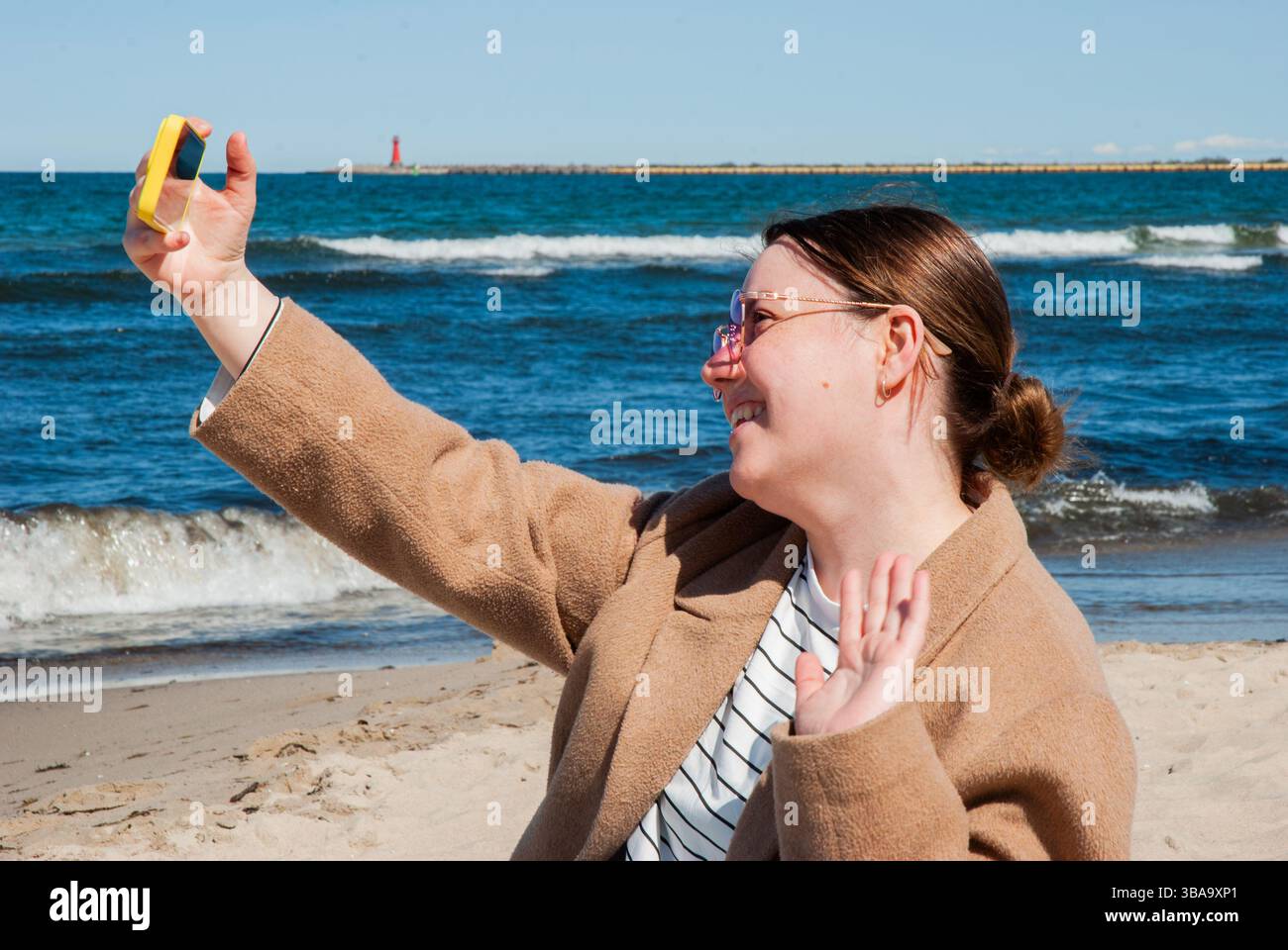 Giovane donna caucasica che scatta un selfie in una giornata di sole sulla spiaggia con le onde e il faro sullo sfondo sotto il cielo azzurro. Nomad digitale Foto Stock