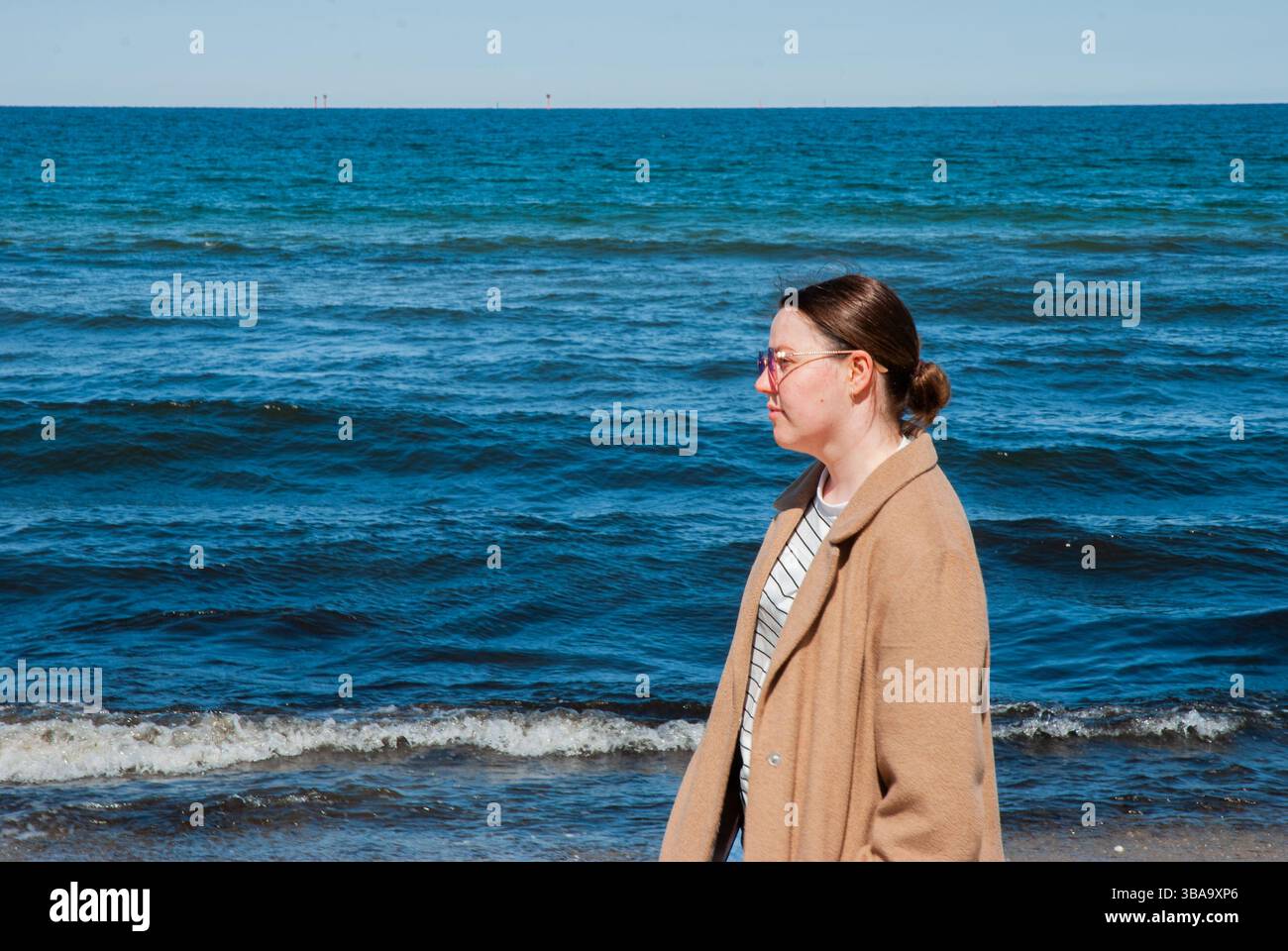 Donna adulta con occhiali che passeggiano sulla spiaggia, serenità delle onde dell'oceano in contrasto con il cielo azzurro per una giornata tranquilla. Nomad digitale Foto Stock