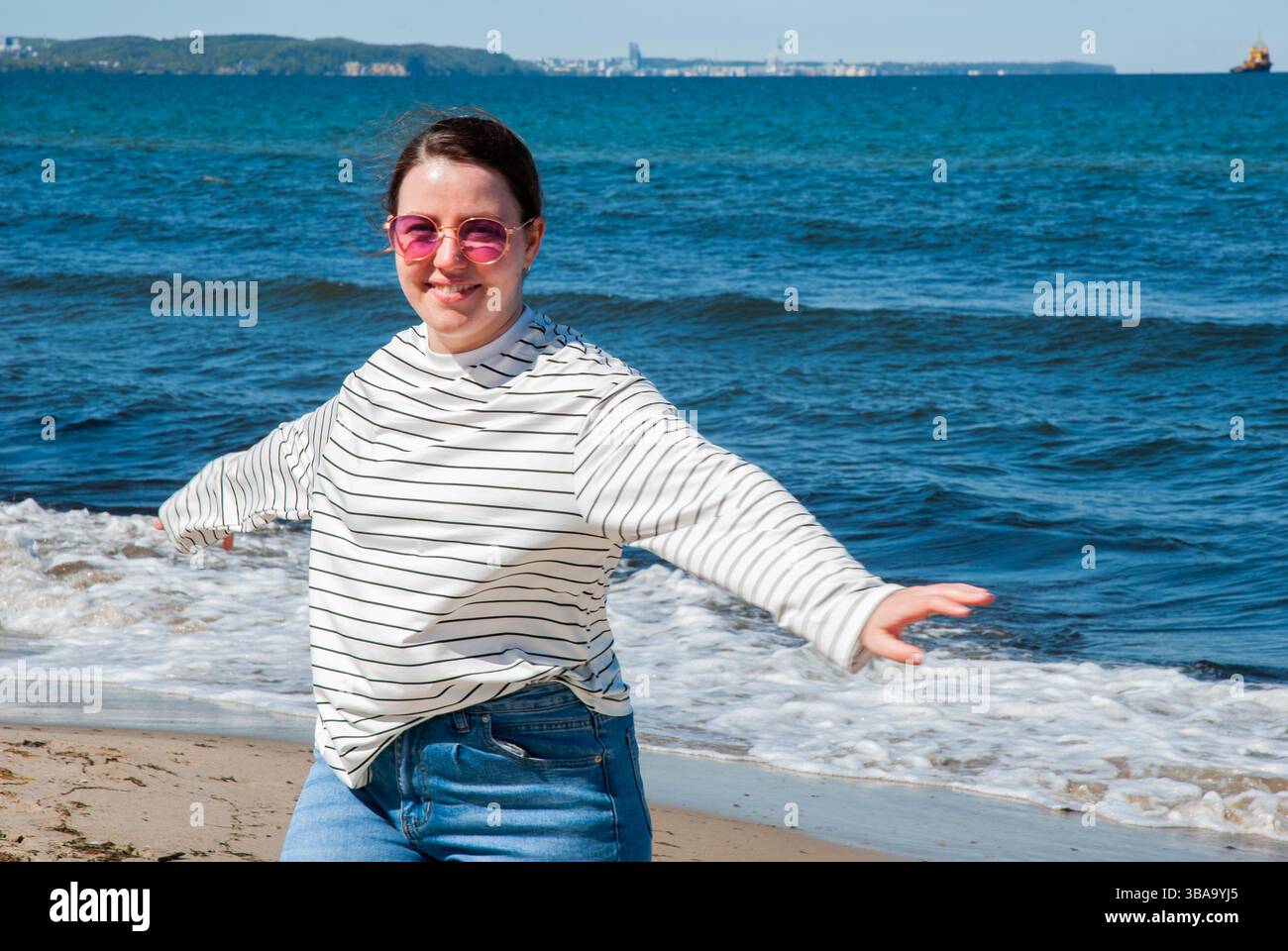 Giovane donna caucasica che si gode una giornata di sole sulla spiaggia, indossando camicia a righe e occhiali da sole, con le braccia allungate e l'oceano sullo sfondo. Cifra Foto Stock