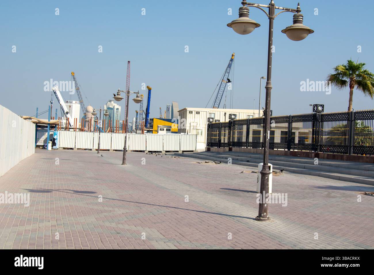 Un cantiere con gru sullo sfondo, caratterizzato da un'ampia passerella vuota fiancheggiata da lampioni e palme. L'area è in fase di sviluppo Foto Stock
