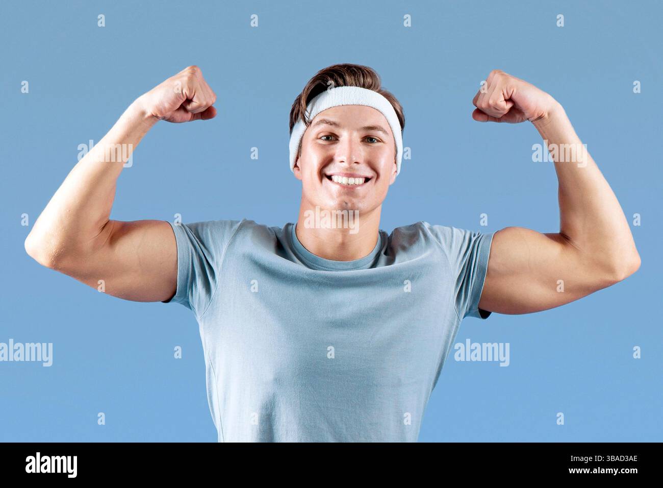 Positivo giovane sportivo seduto su palla fitness e mostrandogli forza su sfondo lilla studio Foto Stock
