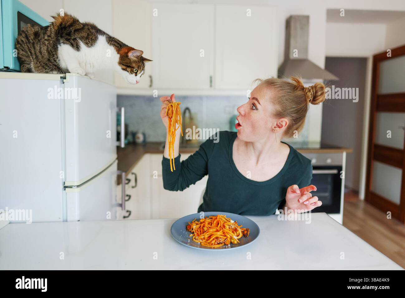 Ragazza che mangia pasta in una cucina accogliente, gatto sul frigorifero aggiunge un'atmosfera curiosa Foto Stock
