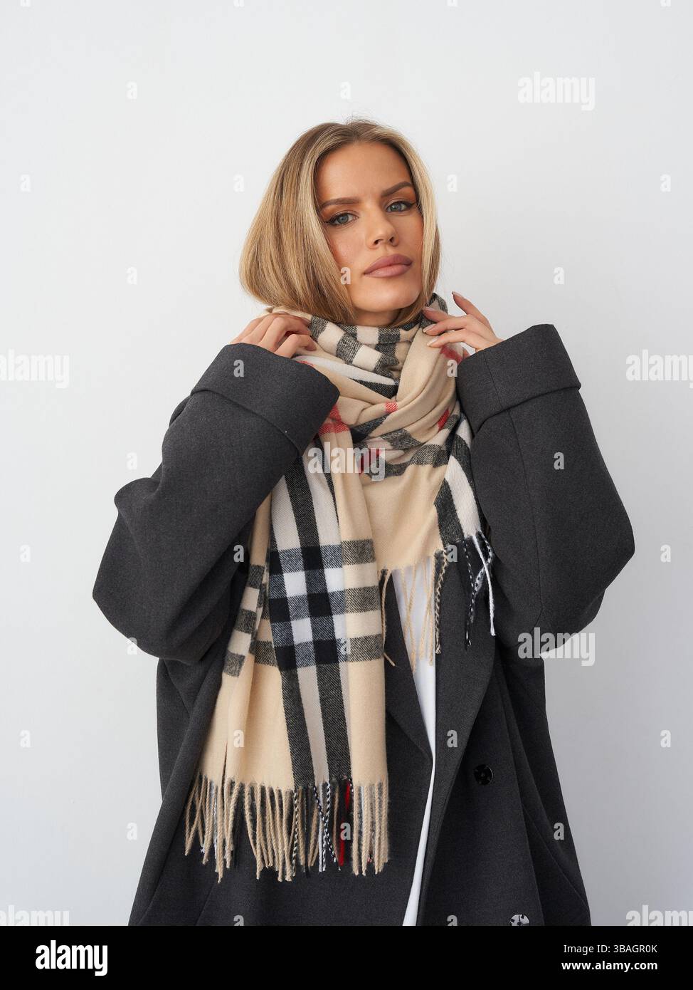 Donna con un lungo cappotto scuro avvolta da una sciarpa a scacchi, che guarda in avanti. I capelli marrone chiaro sono modellati in modo elegante. La sciarpa presenta nero, rosso e beige Foto Stock