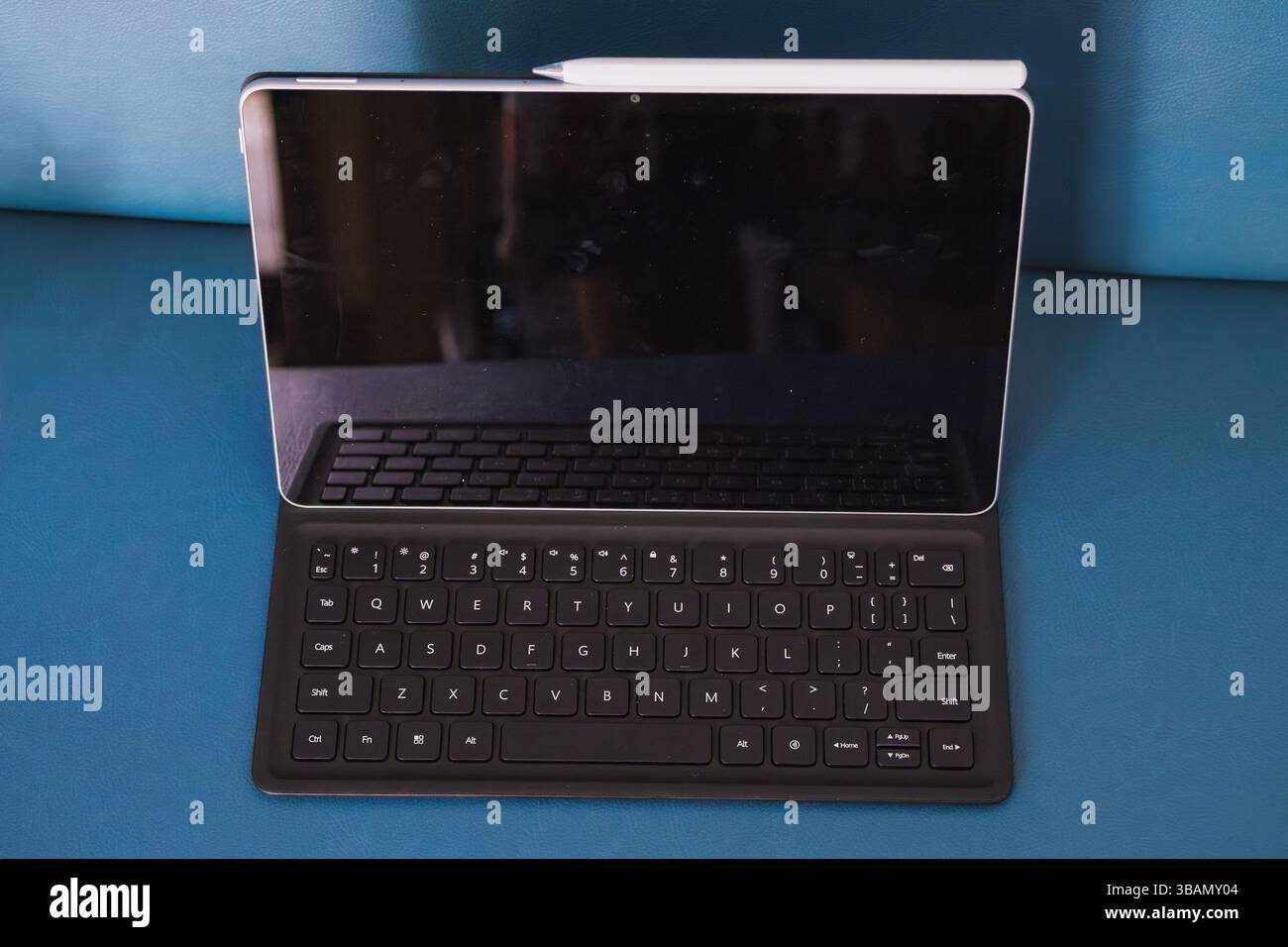 Un tablet PC con una tastiera nera e uno stilo bianco si appoggia su una superficie blu testurizzata. Lo schermo è scuro e riflette l'ambiente circostante. Foto Stock