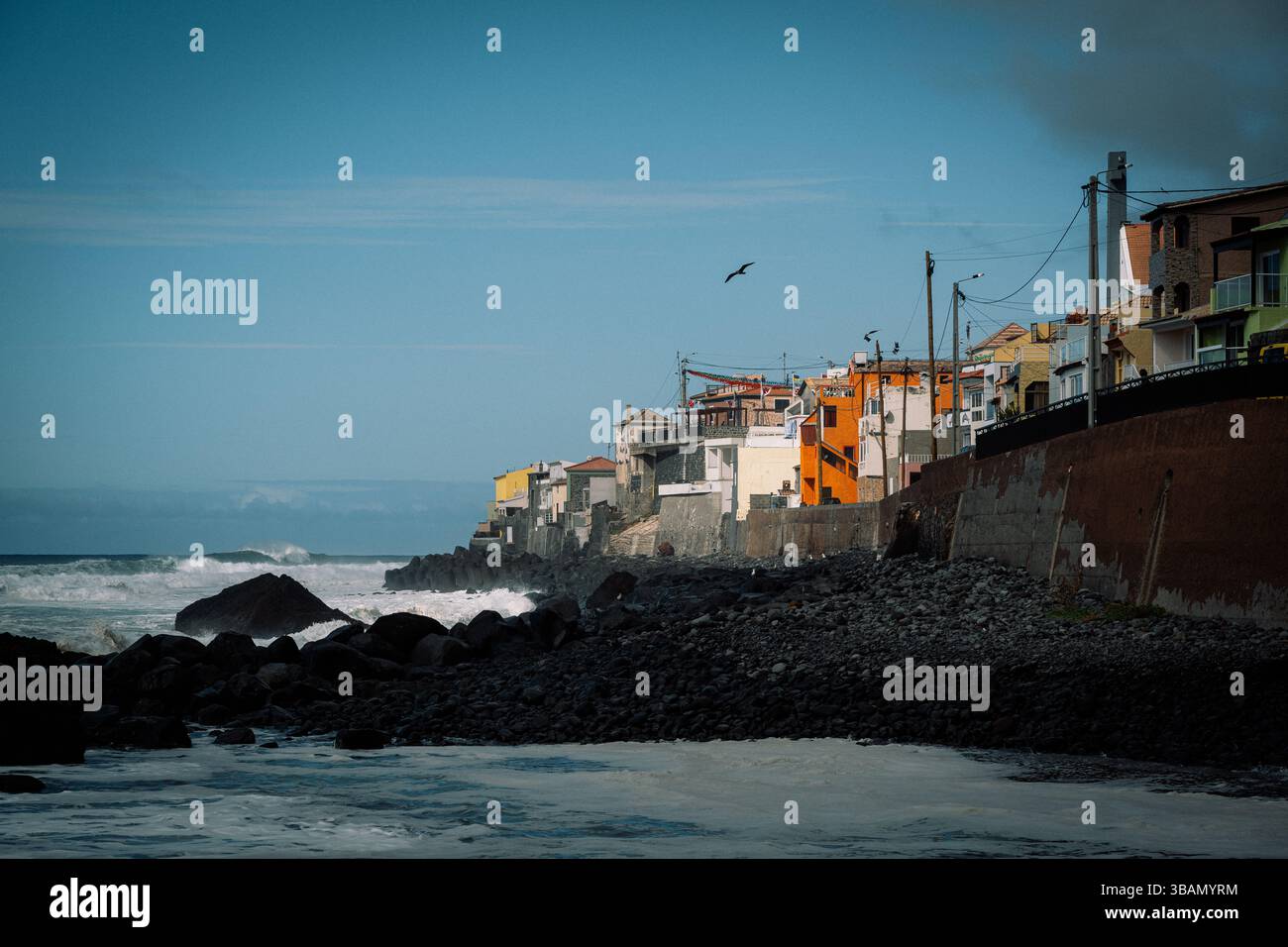Colorato villaggio costiero arroccato sulla costa rocciosa con onde che si infrangono sotto un cielo blu Foto Stock