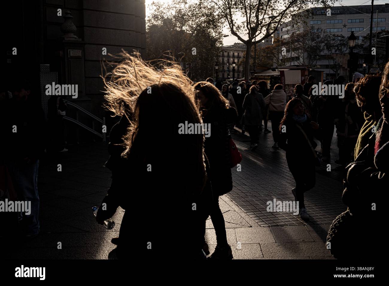 Persone che camminano attraverso una strada della città di Barcellona, in Spagna, in una giornata molto ventosa, con raffiche forti, silhouette retroilluminate e luce del sole pomeridiano. Foto Stock