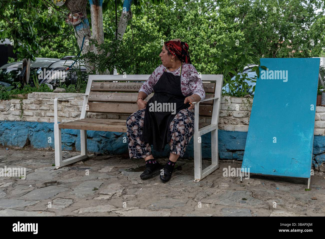A Sirince, un'anziana donna in abito a fantasia siosa siede su una panchina bianca accanto a una porta blu, circondata da lussureggiante vegetazione, che evoca un villaggio rurale turco Foto Stock